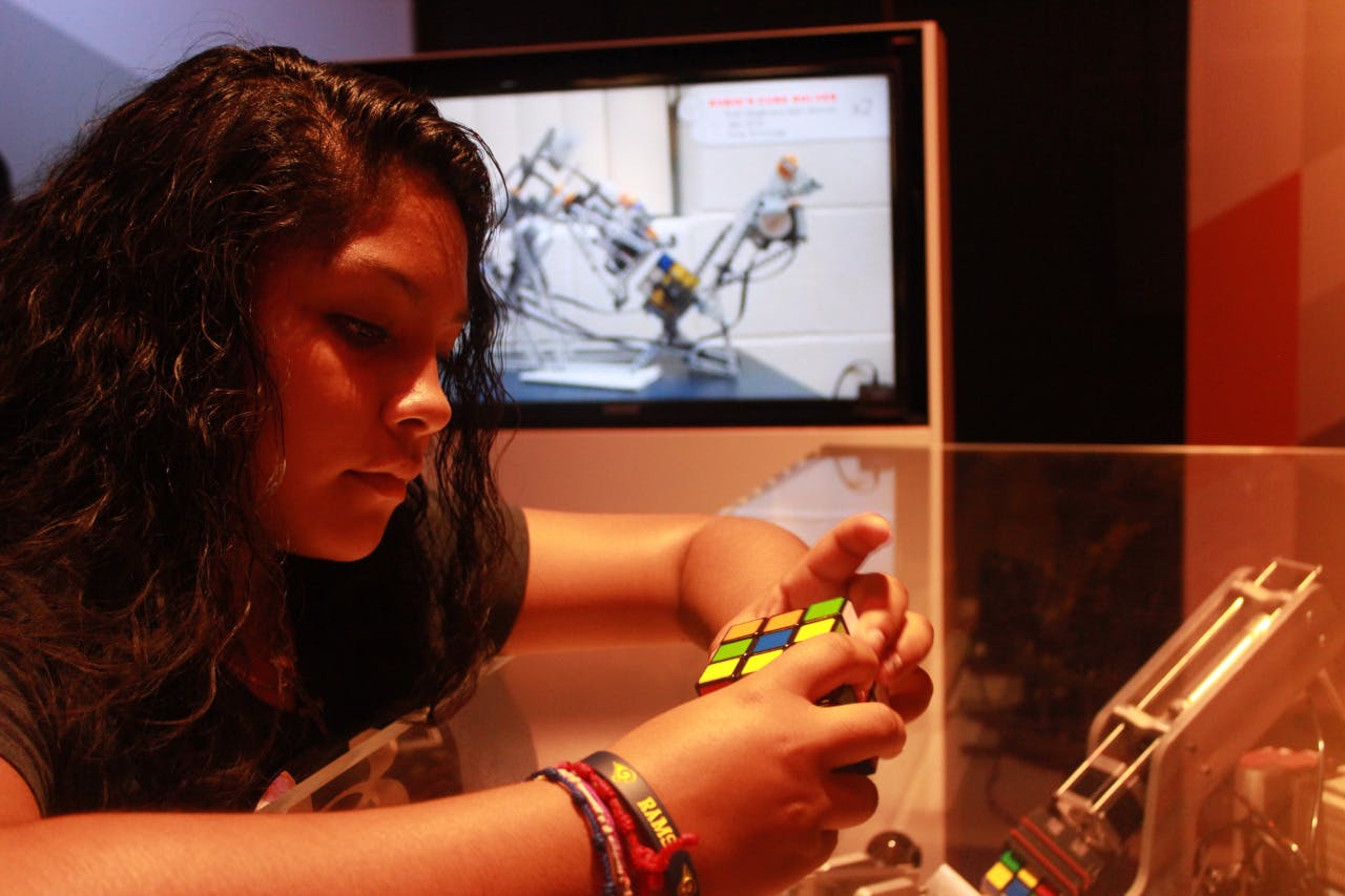 Person solving a Rubik's Cube with a screen displaying a robot in the background