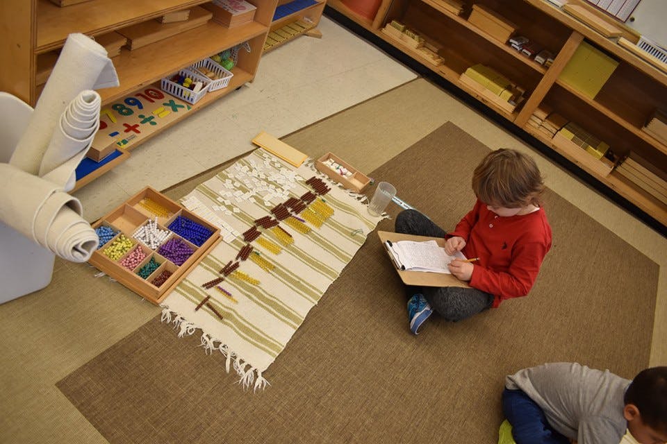 Child sitting on floor writing on a clipboard next to a rug with objects
