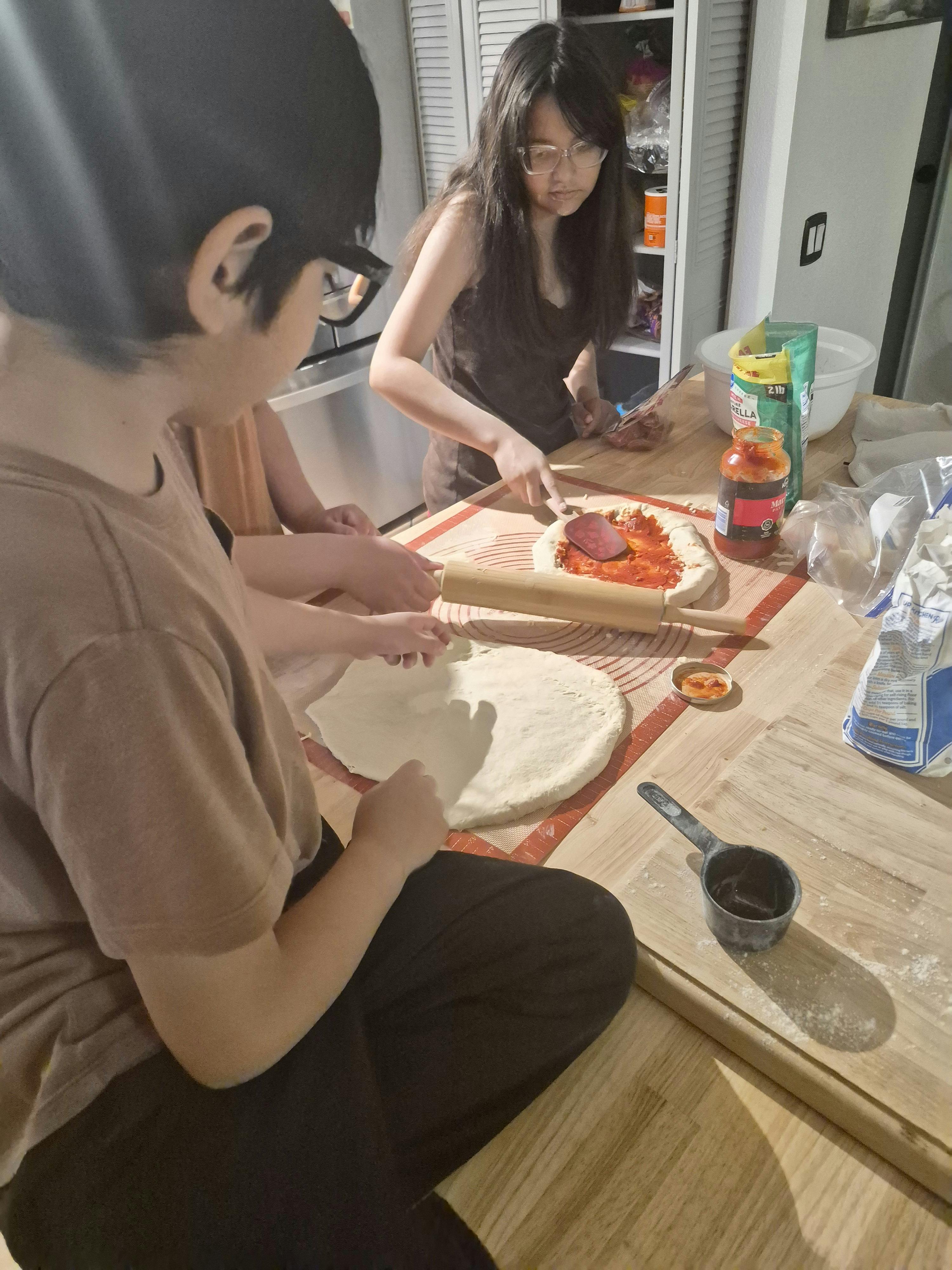 People rolling out dough and spreading sauce to make pizza in a kitchen