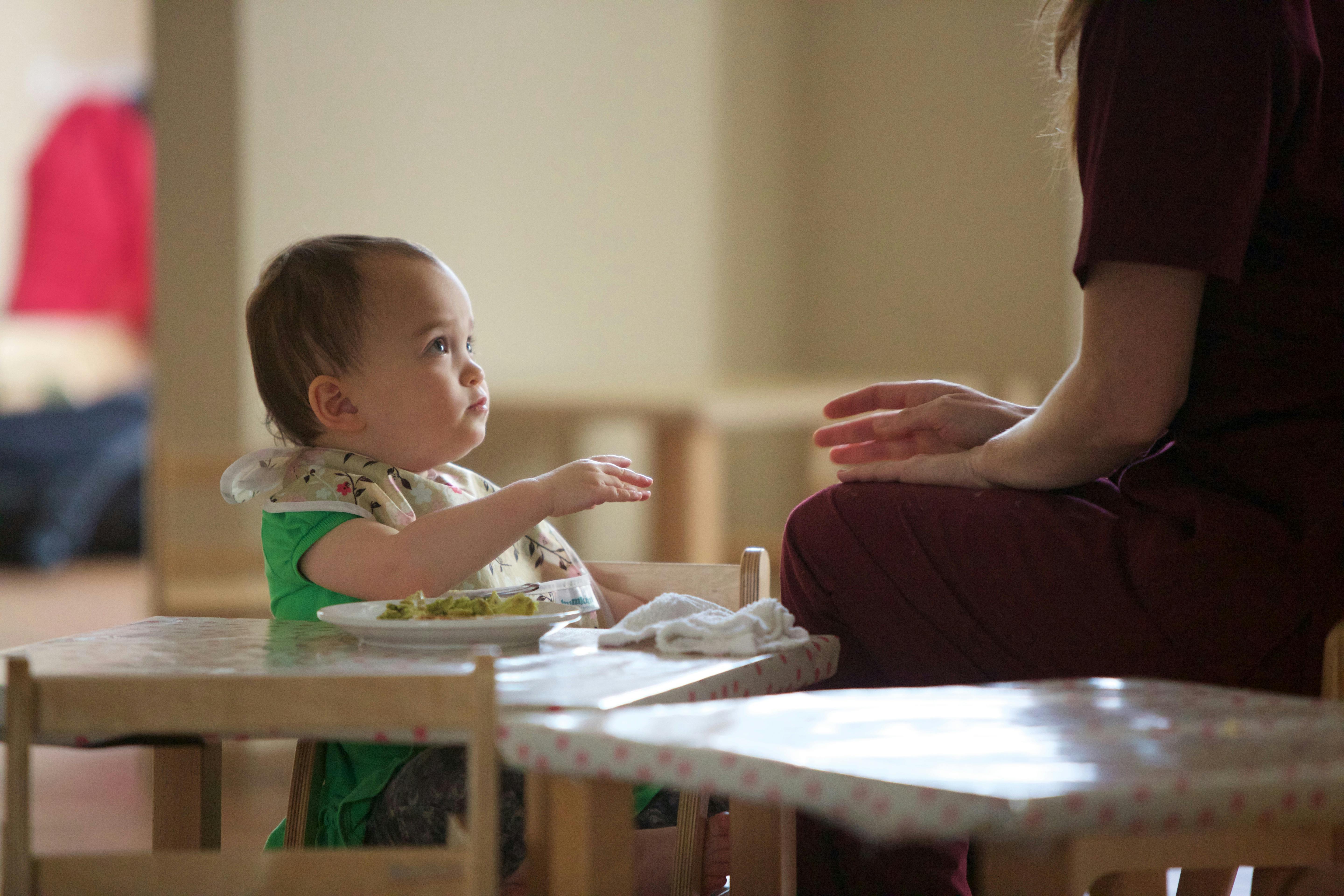 Child in a bib sitting at a table with a plate of food, facing a person