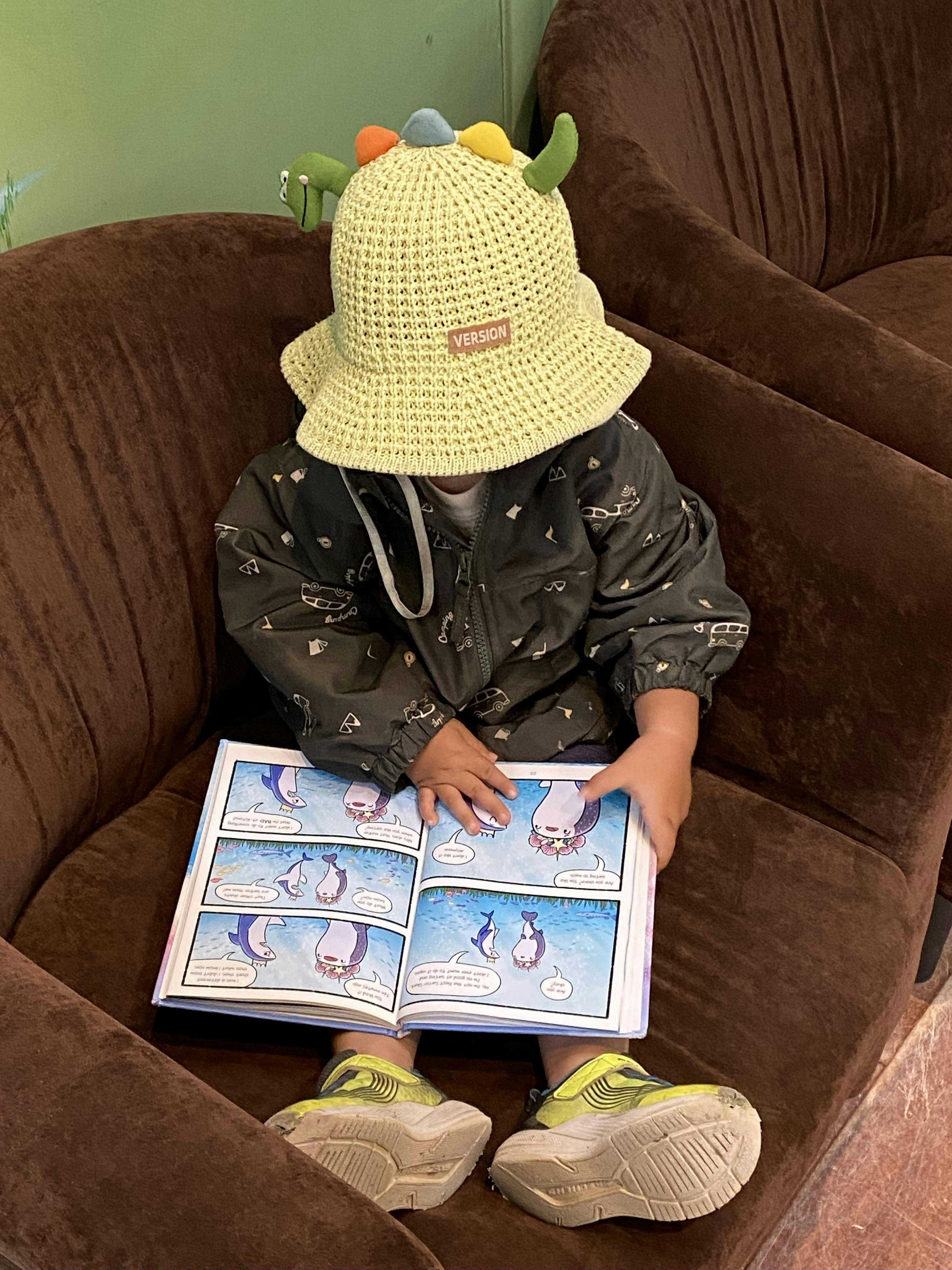 Child wearing a dinosaur hat reading a comic book on a couch