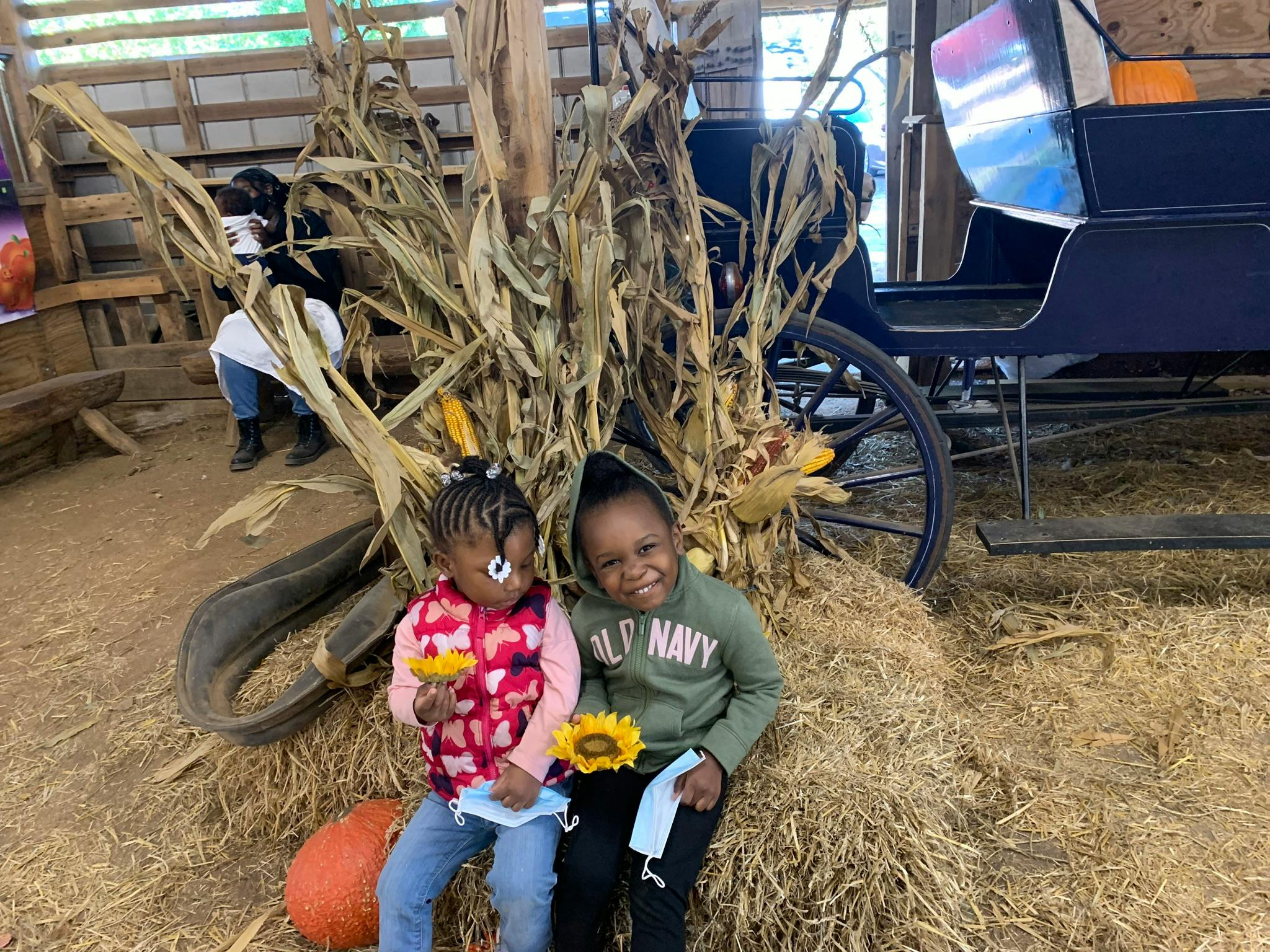 Two children sitting on hay bales with corn stalks and a carriage