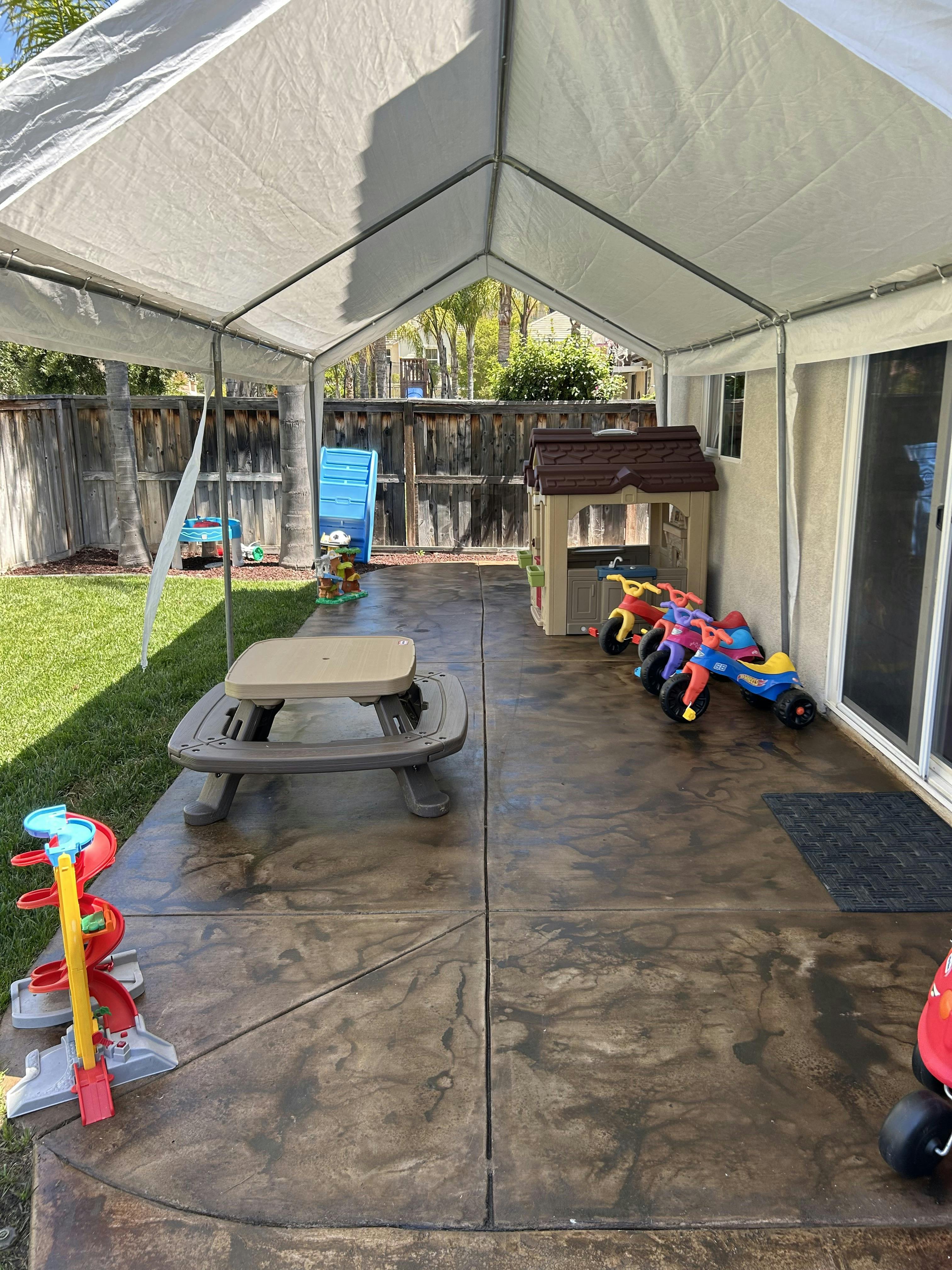 A canopy covers an outdoor play area with a picnic table, playhouse, and ride-on toys