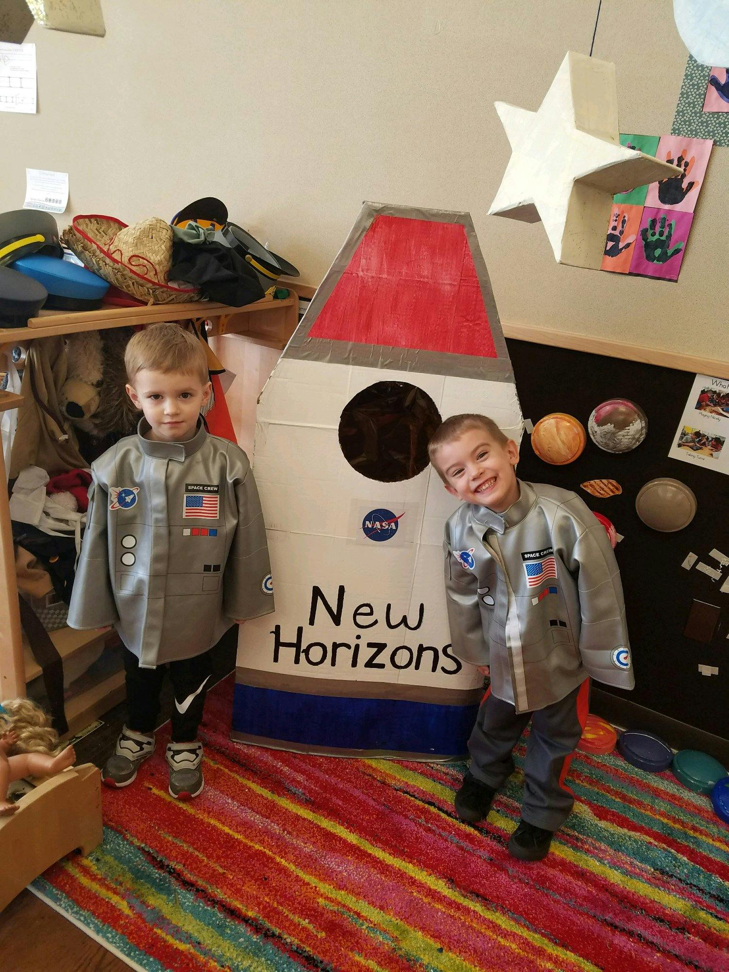 Two children in astronaut costumes standing next to a cardboard rocket labeled "New Horizons"