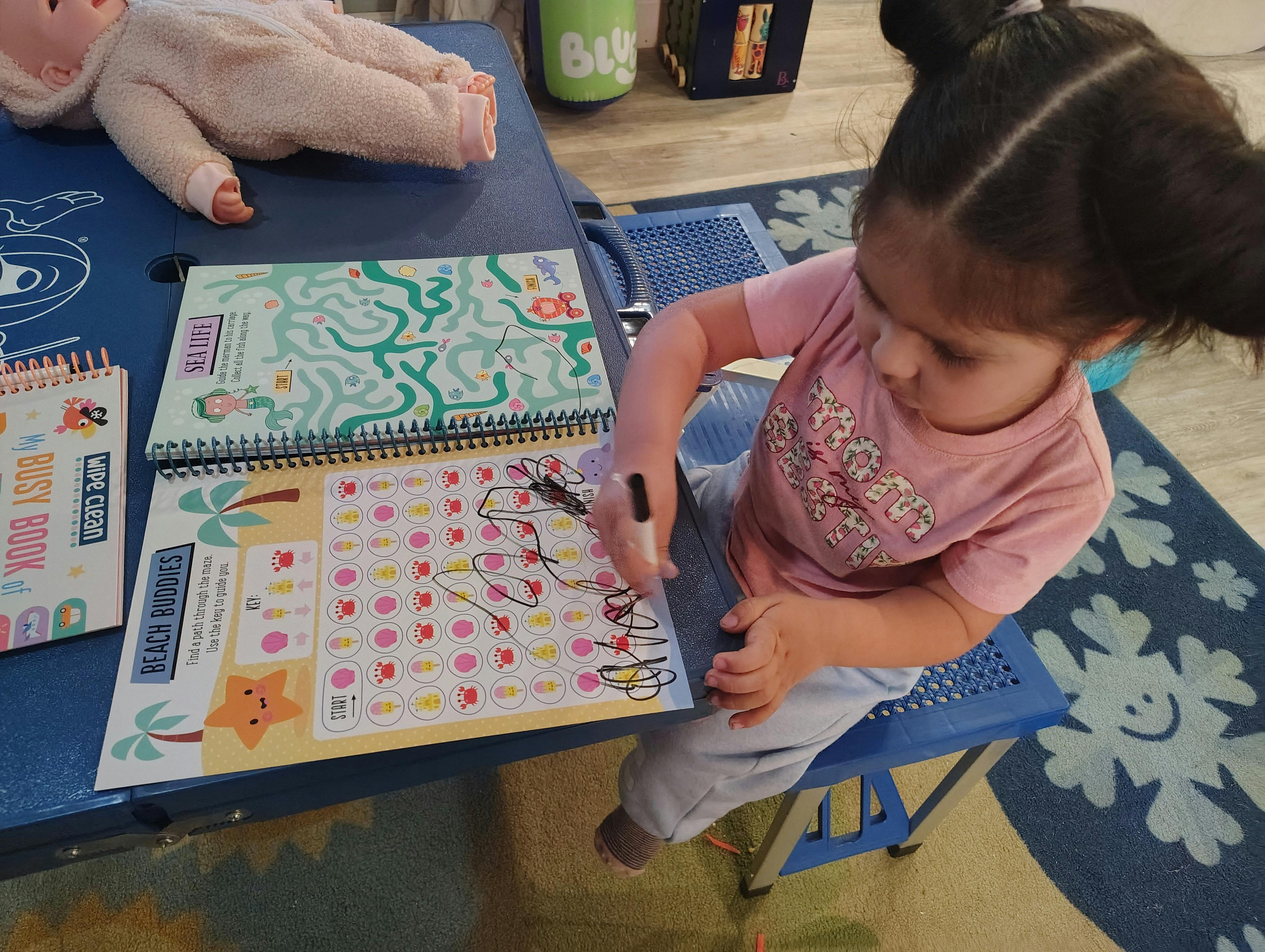 Child drawing on a 'Beach Buddies' activity book with a doll nearby