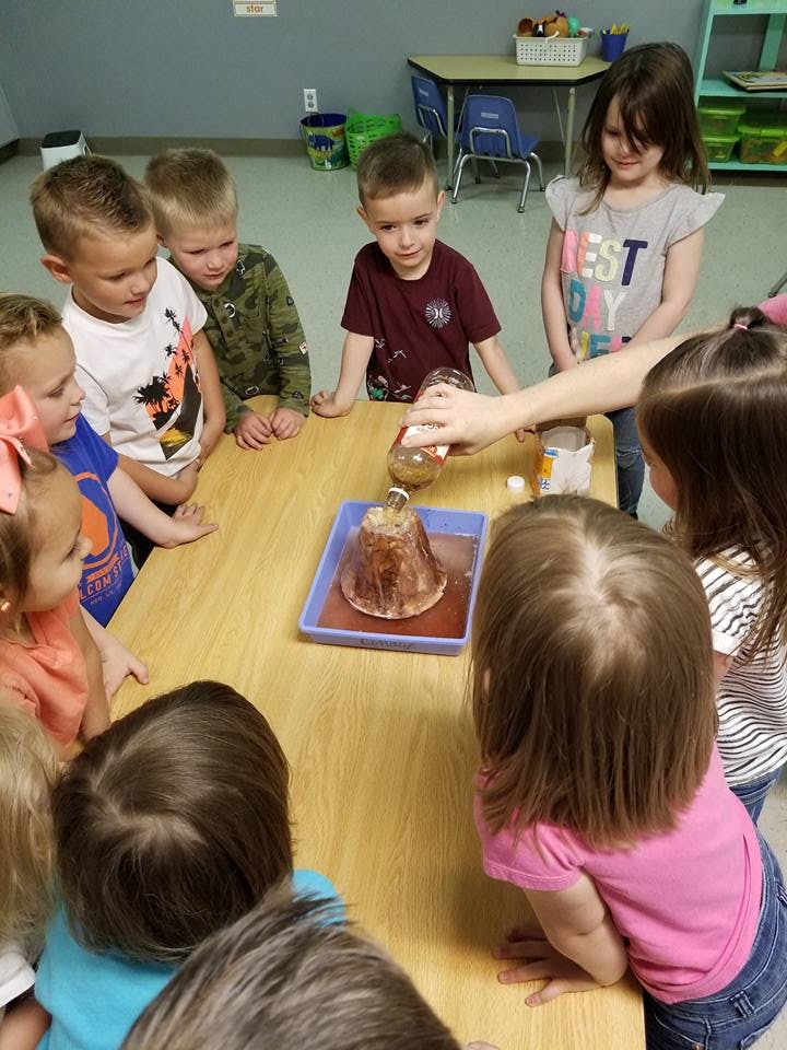 Children gathered around a table watching a person pour liquid into a volcano model