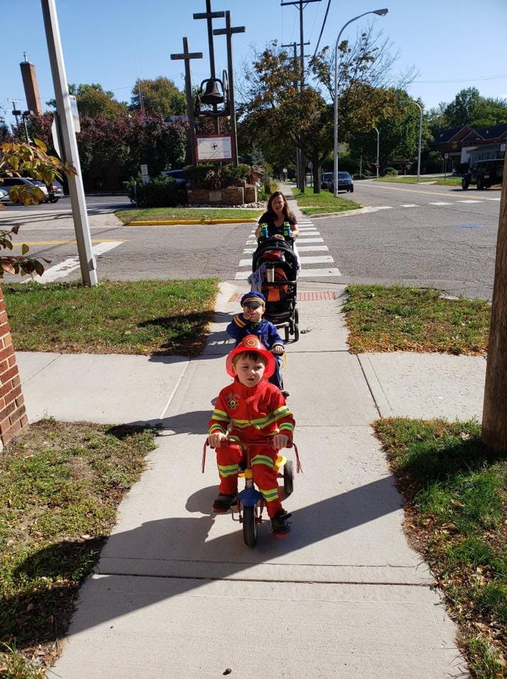Child in firefighter costume on tricycle, child in stroller, person pushing stroller