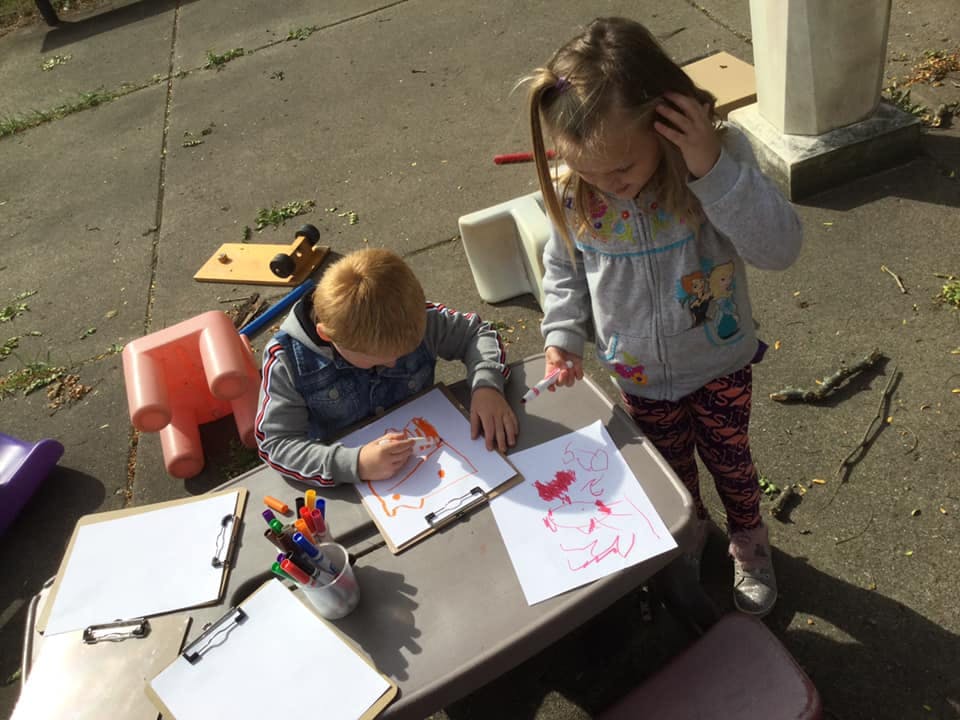 Child drawing at a table while another child stands nearby