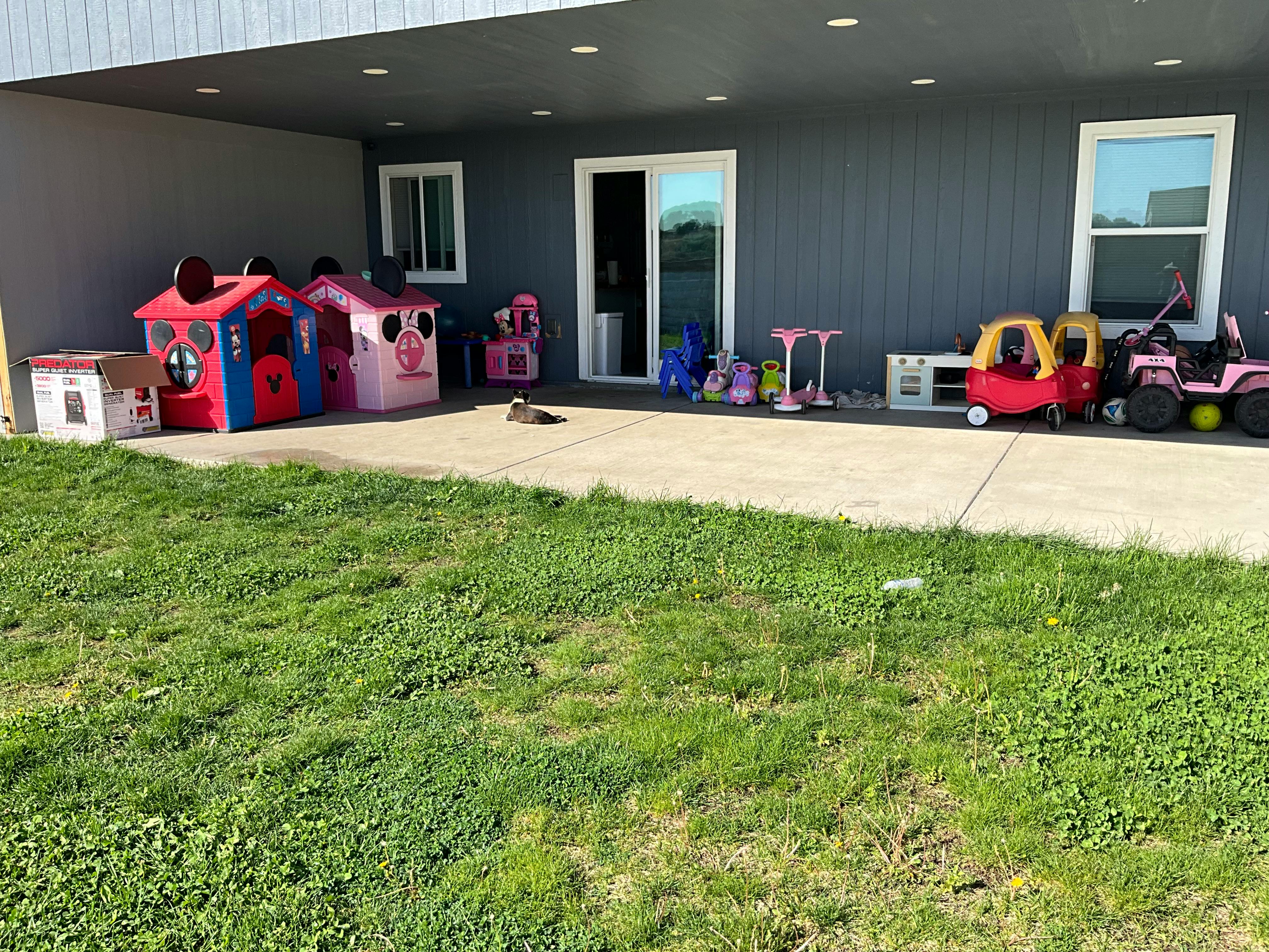 Playhouses and ride-on toys on a patio next to a house with a grass lawn