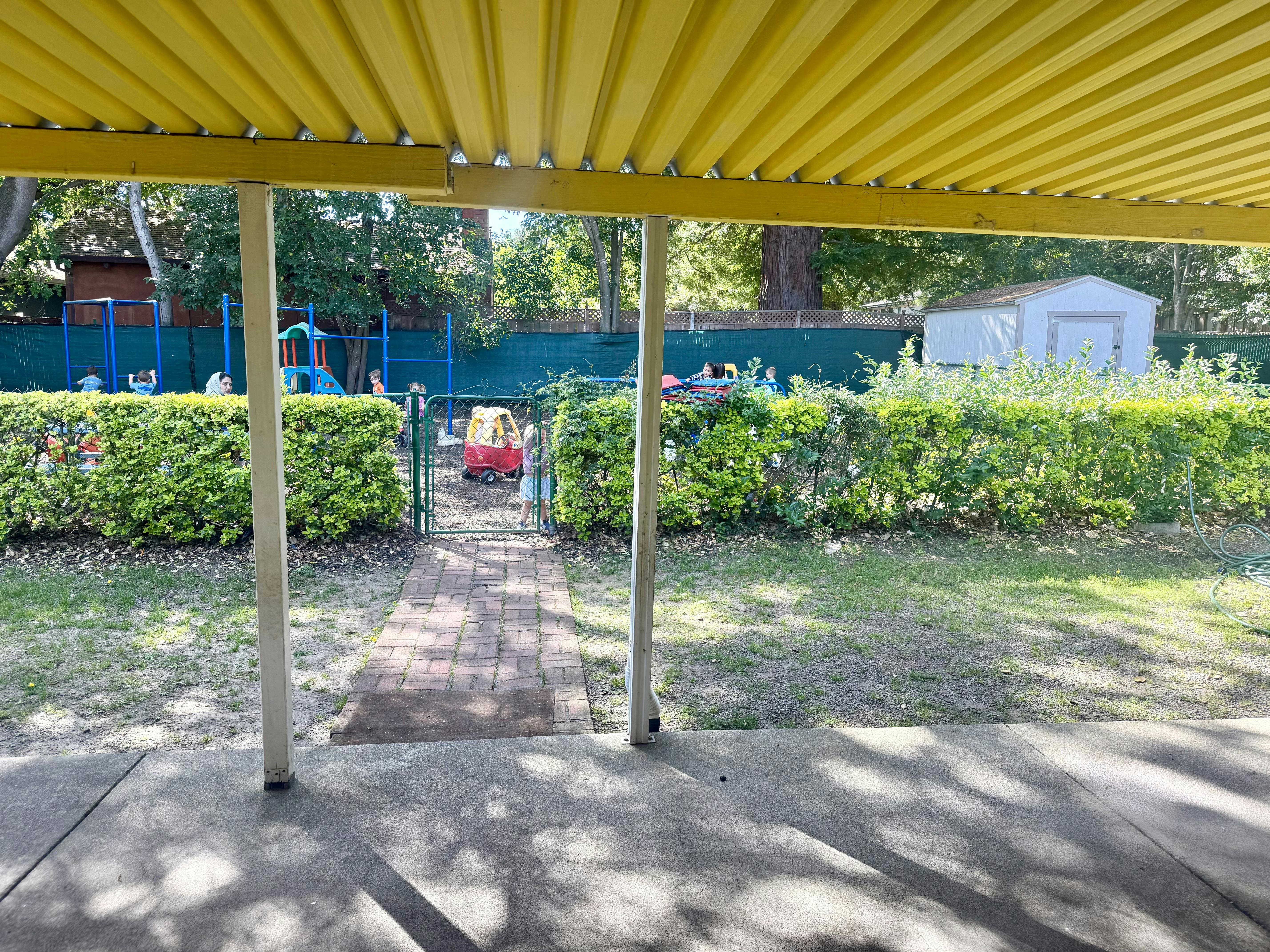 Children playing in a fenced outdoor playground seen from under a patio cover