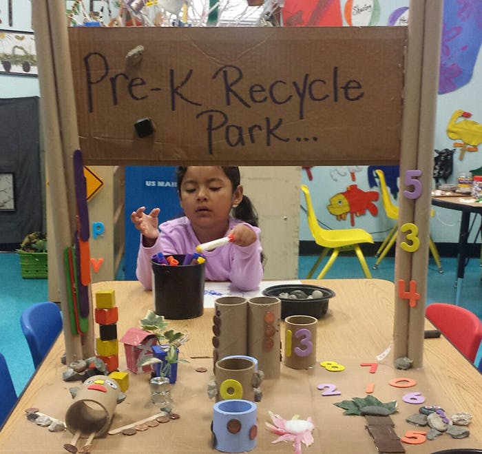 Child at a table with craft supplies under a sign that reads "Pre-K Recycle Park"