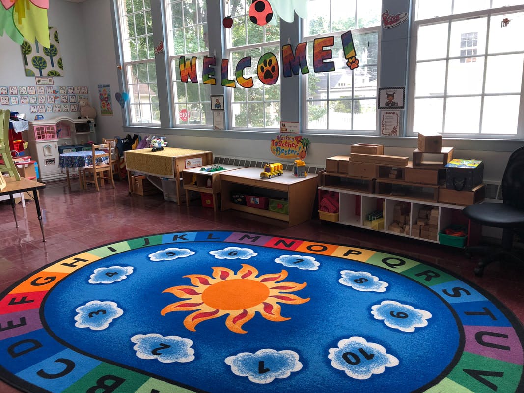 Classroom with a colorful rug featuring a sun and clouds, a "WELCOME!" banner