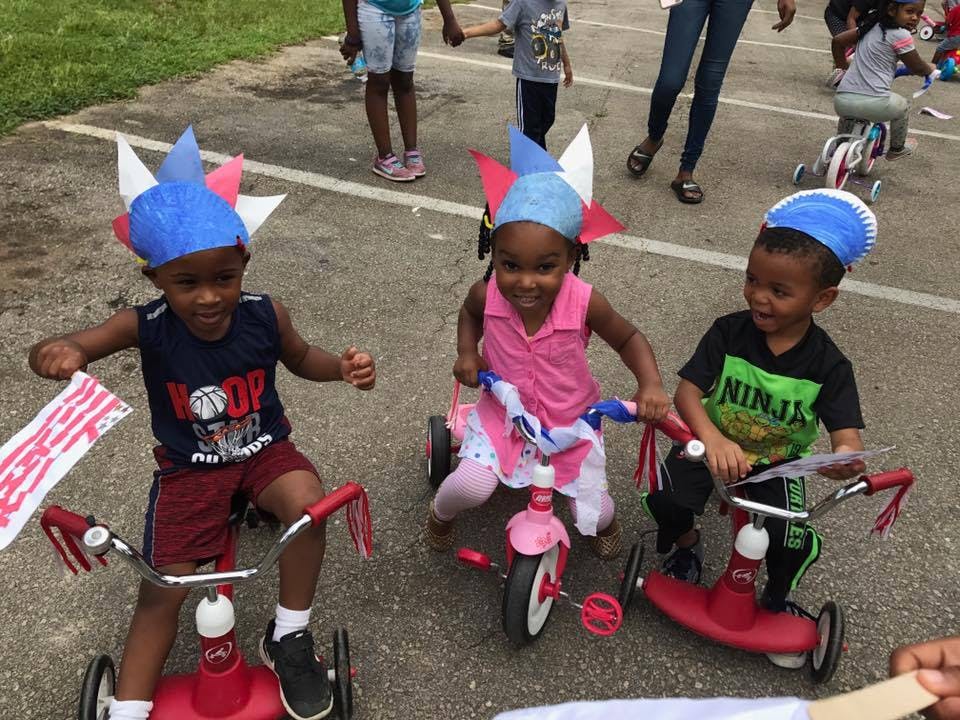 Three children on tricycles wearing patriotic hats