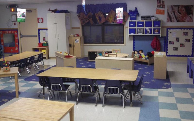 Empty classroom with small tables and chairs, a play area with dolls, and storage shelves
