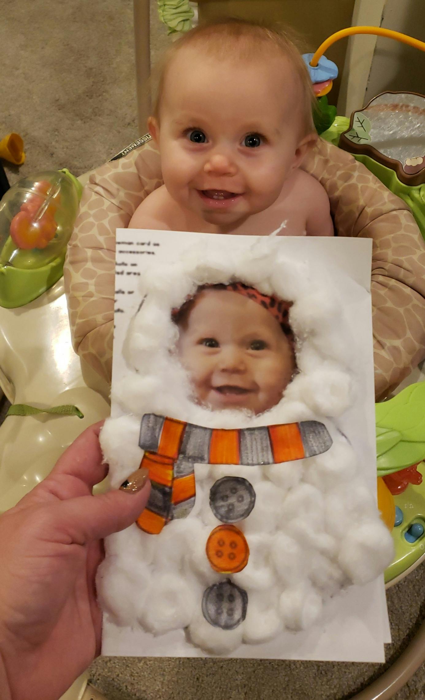 Child in a bouncer with a person holding a snowman craft with a child's face