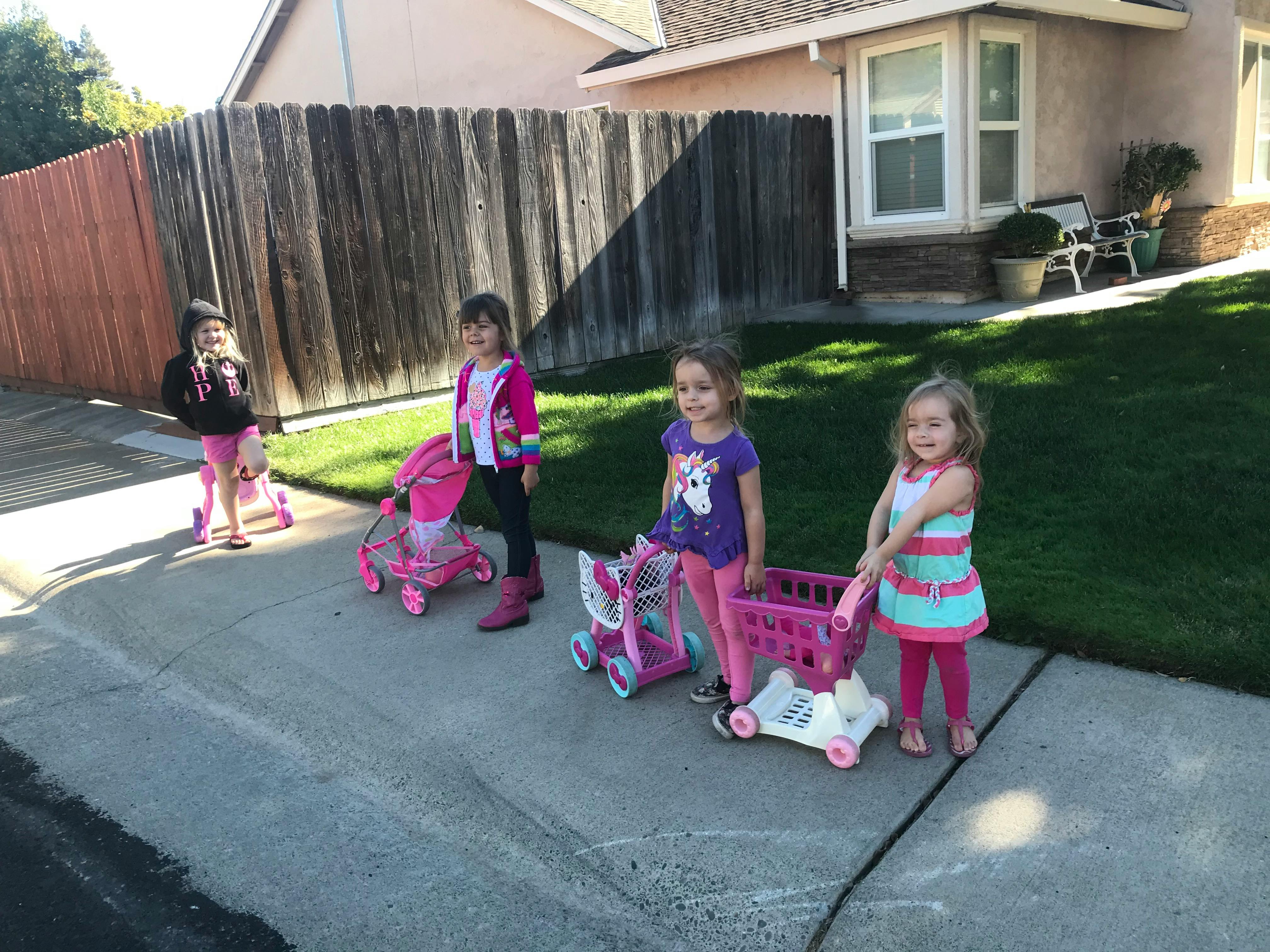 Four children playing with toy strollers and shopping carts on a sidewalk