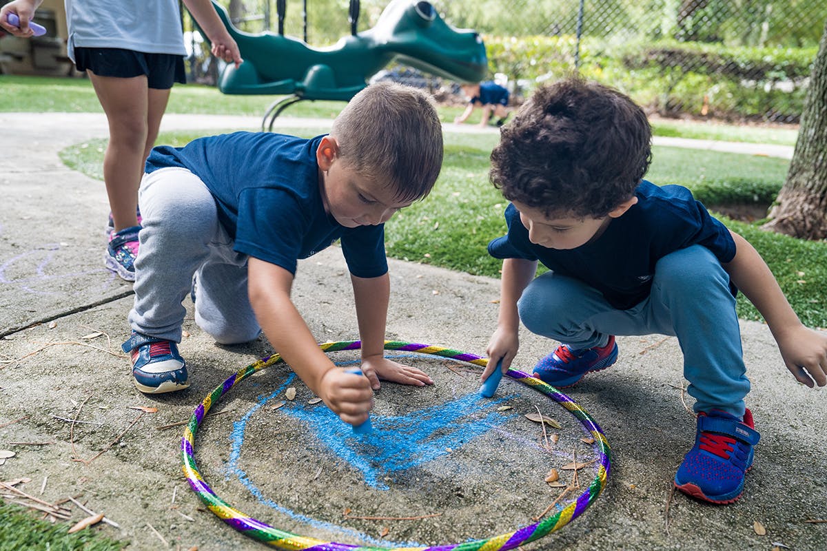 Two children drawing with chalk inside a hula hoop on the ground