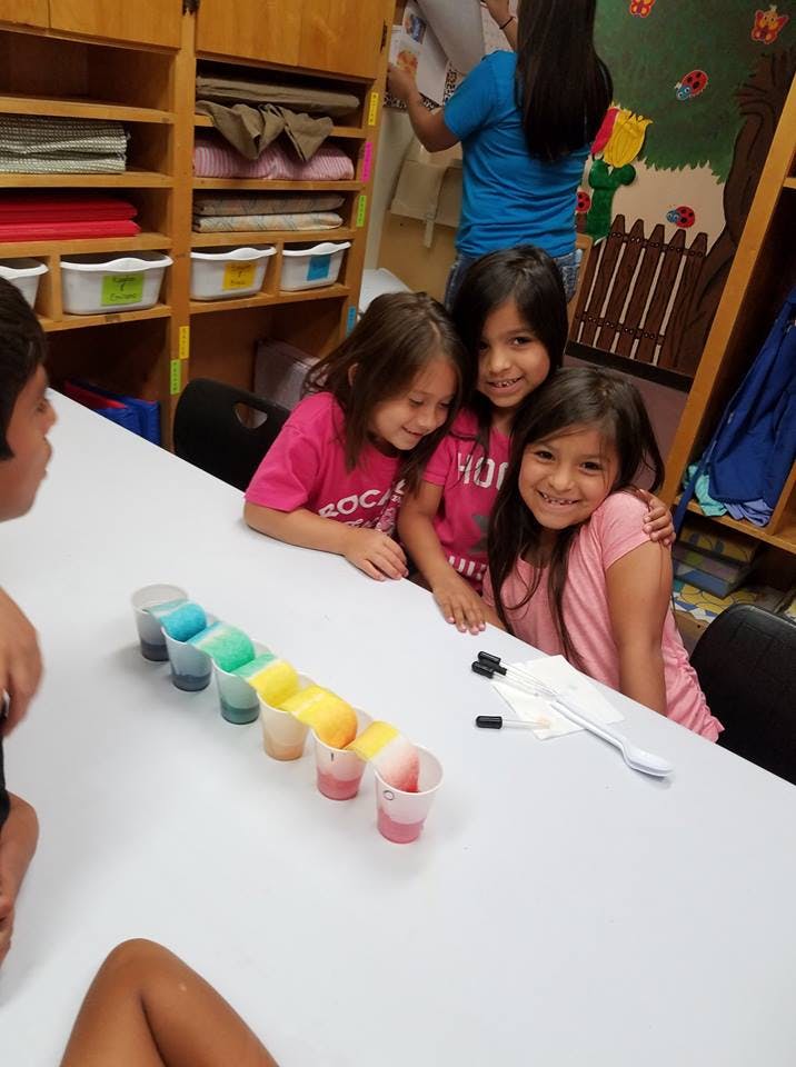 Children at a table with cups of colored liquid and clear liquid connected by paper towels