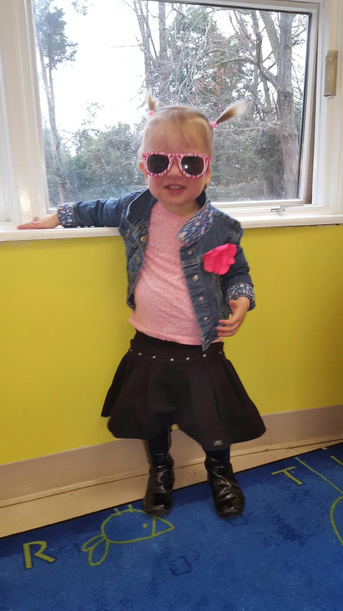 Child in sunglasses, denim jacket, and black skirt standing by a window