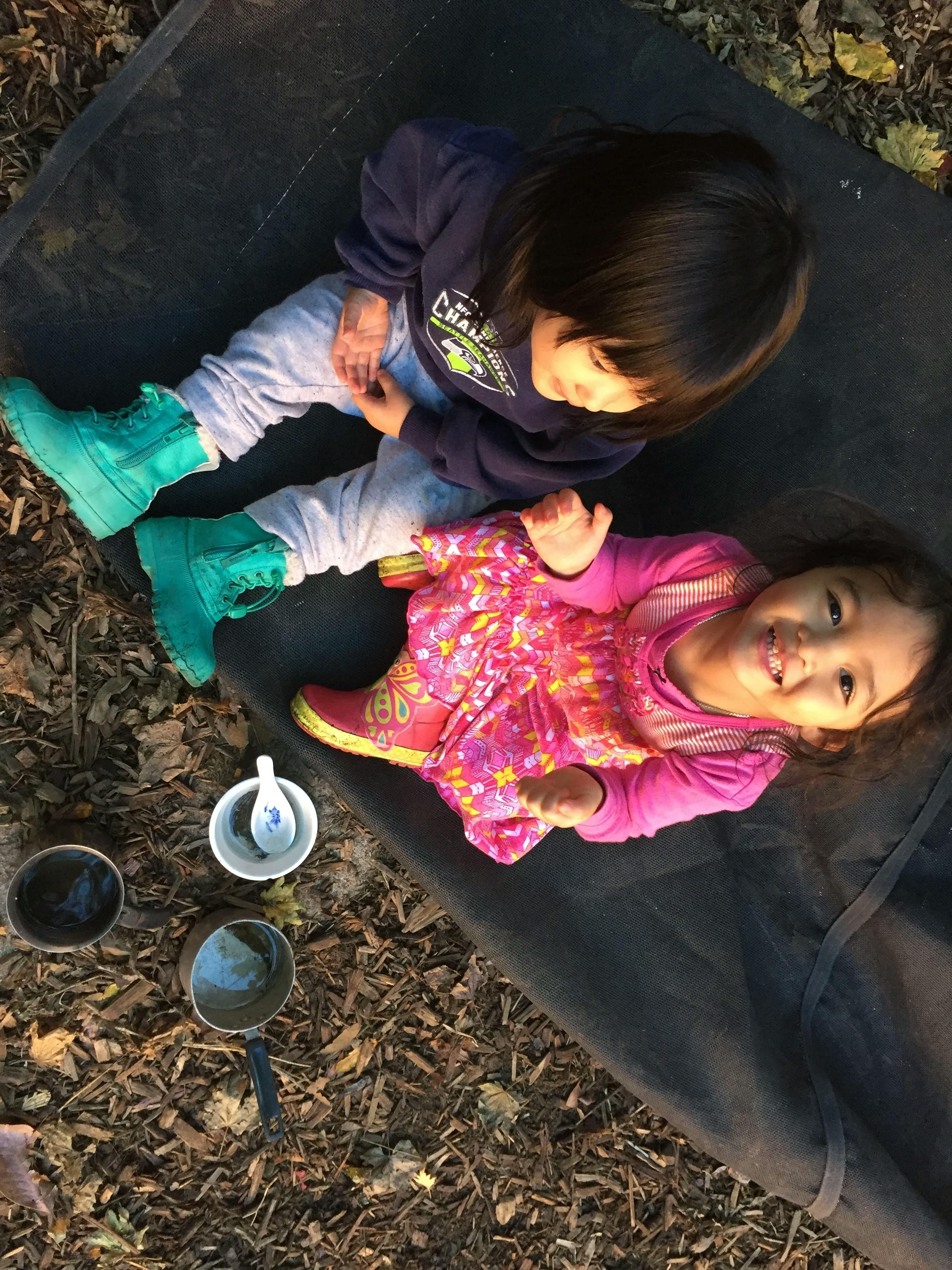 Two children sitting on a mat with small pots and cups around them