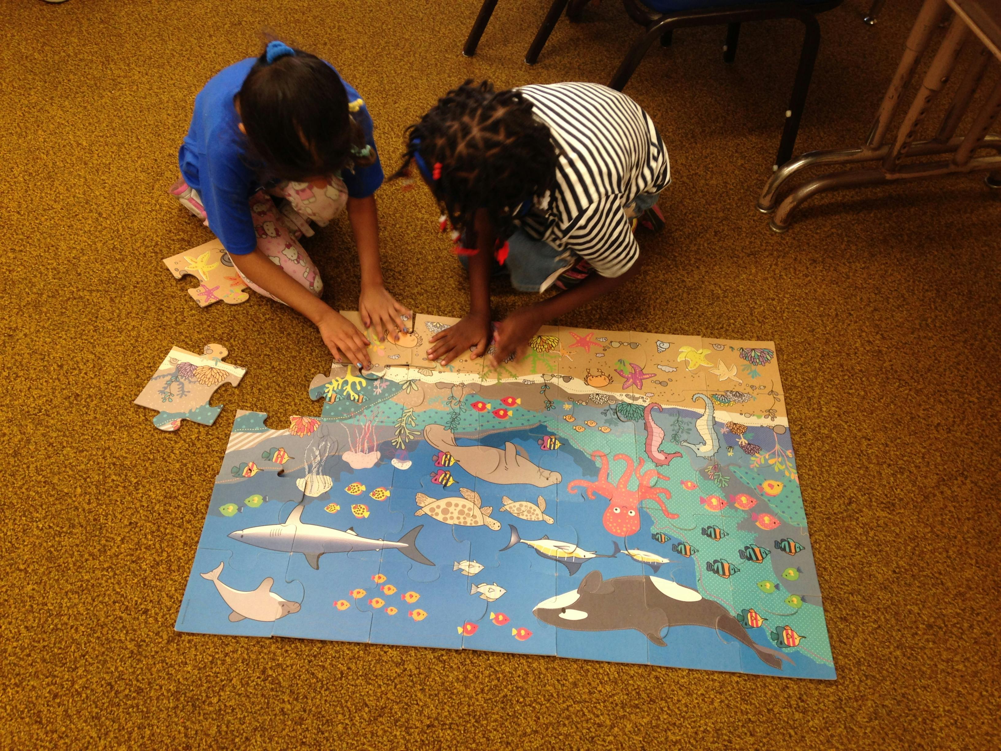 Two children kneeling on the floor assembling a large ocean-themed puzzle