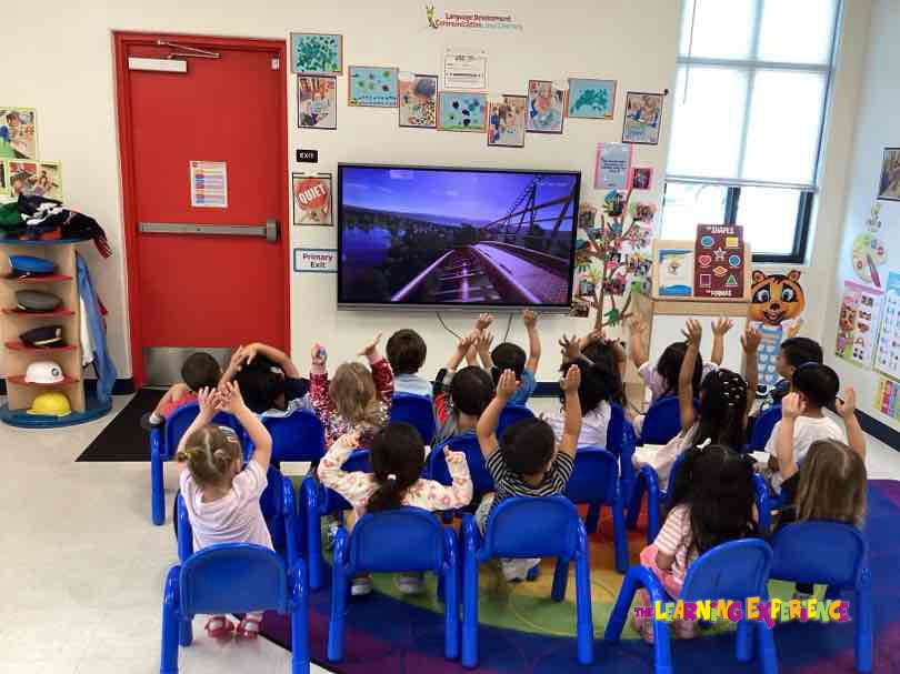 Children sitting in blue chairs with their hands raised toward a TV in a classroom