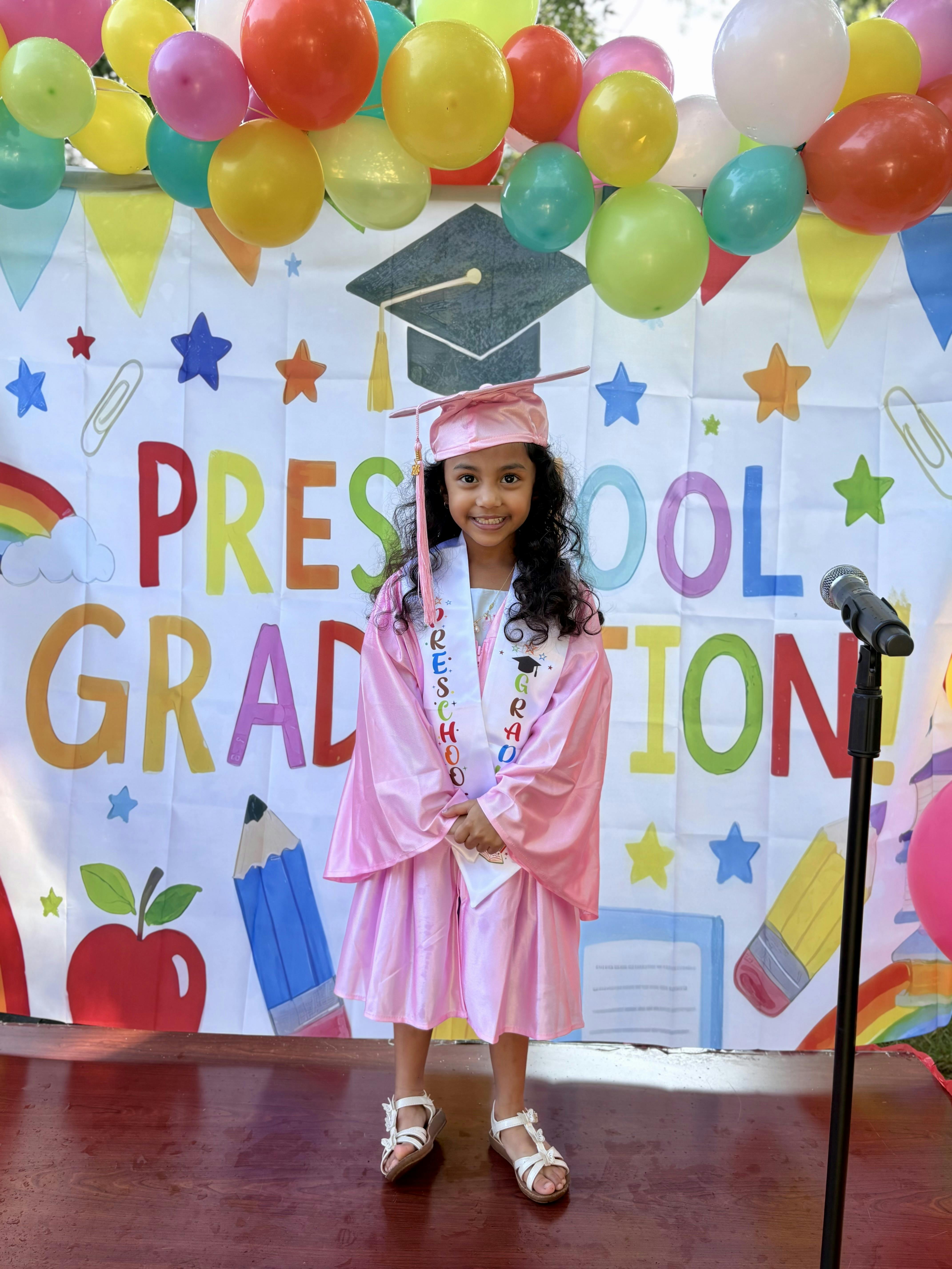Child in pink graduation cap and gown standing in front of a backdrop