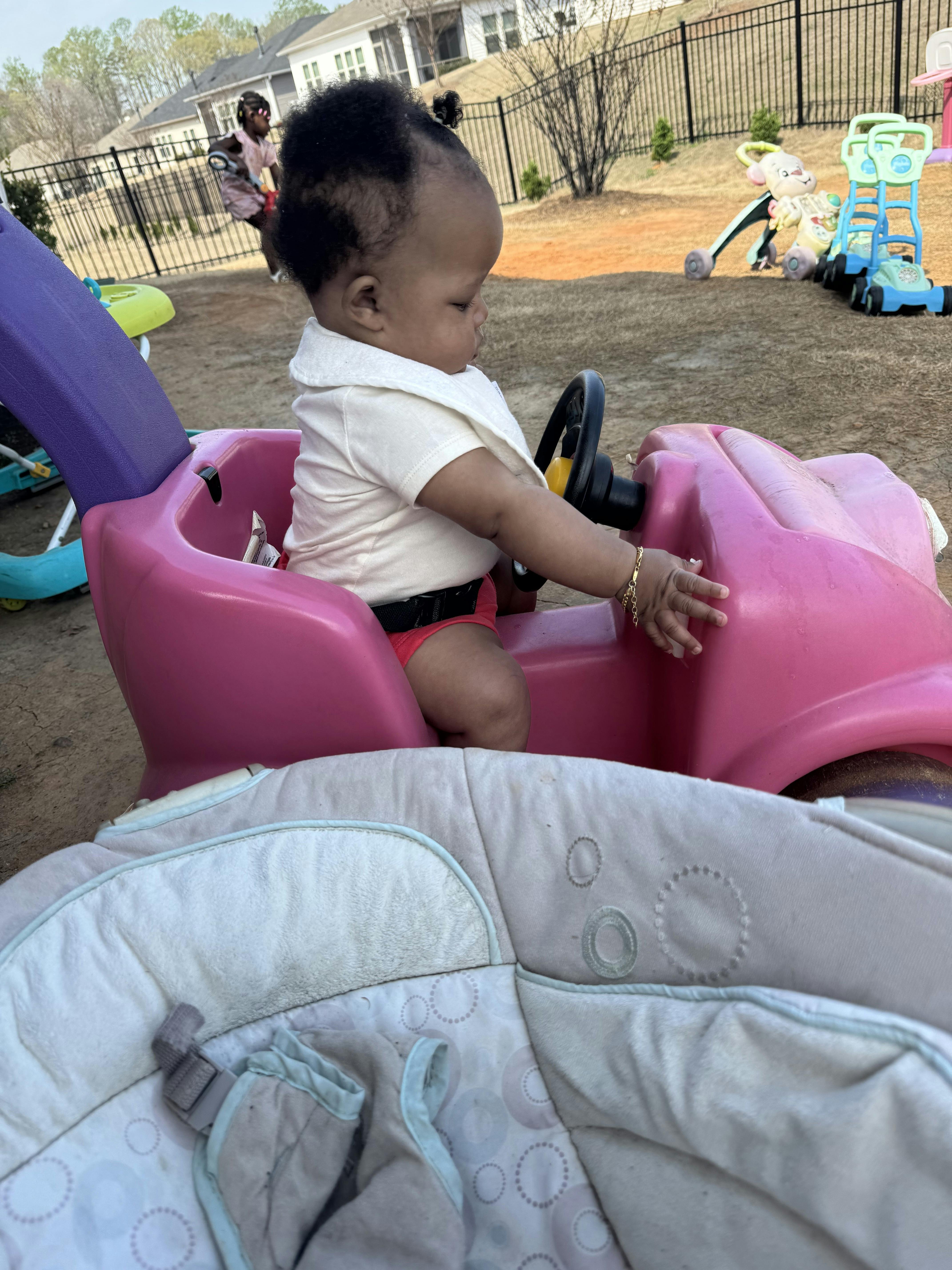 Child sitting in a pink toy car with hands on the steering wheel