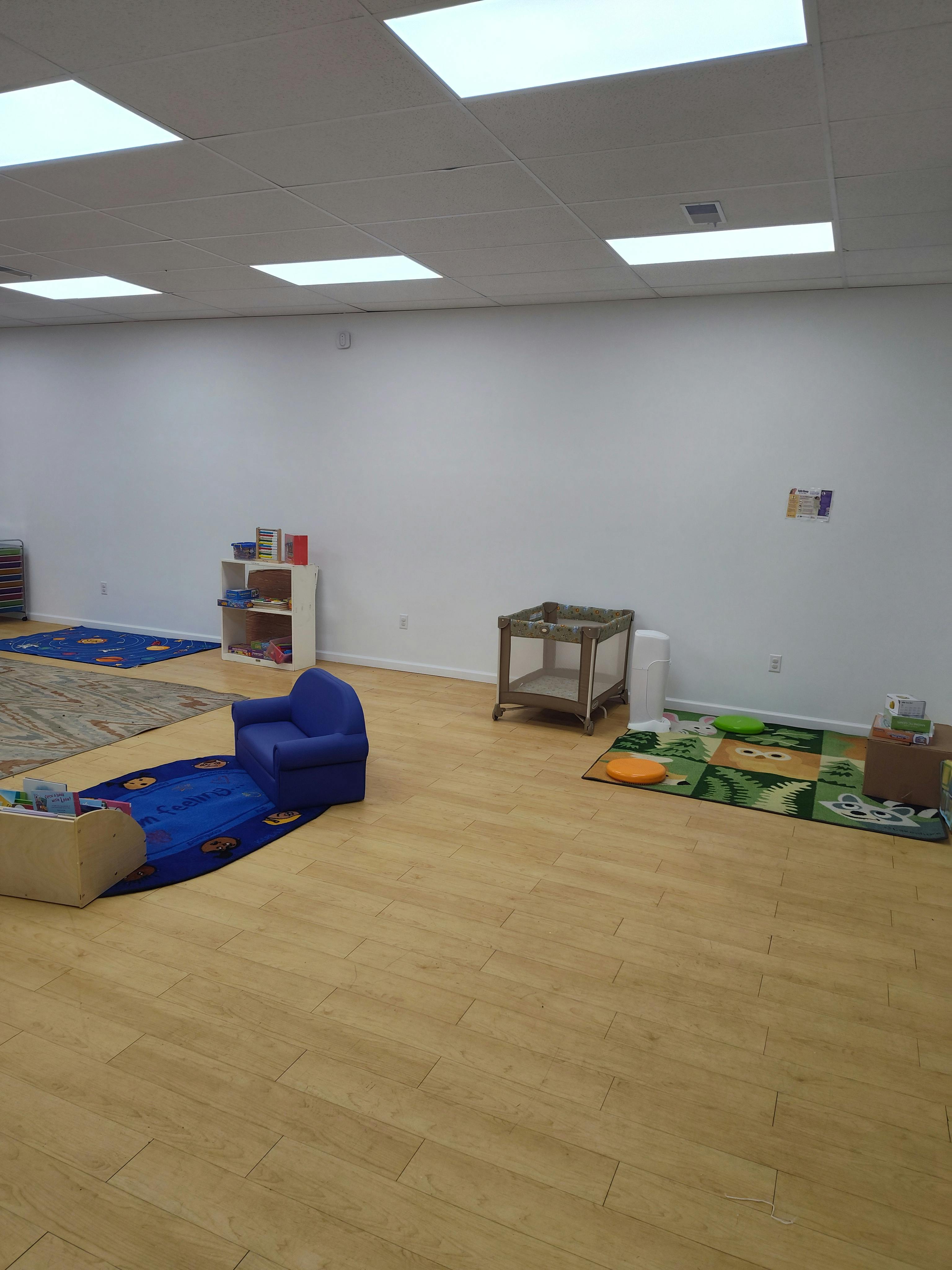 Empty classroom with small blue chair, playpen, and rugs on a wooden floor