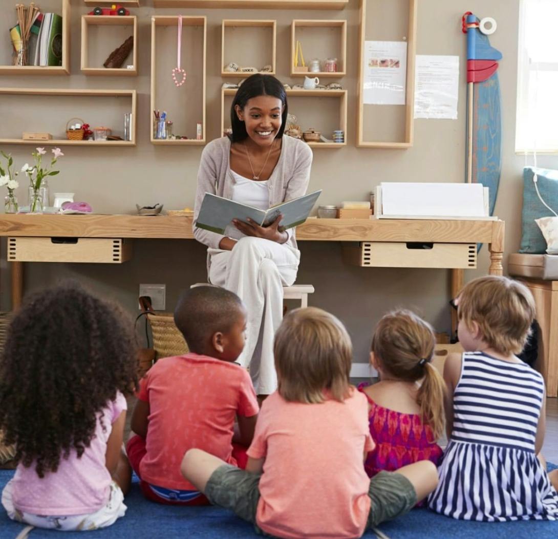 Person reading a book to a group of children sitting children