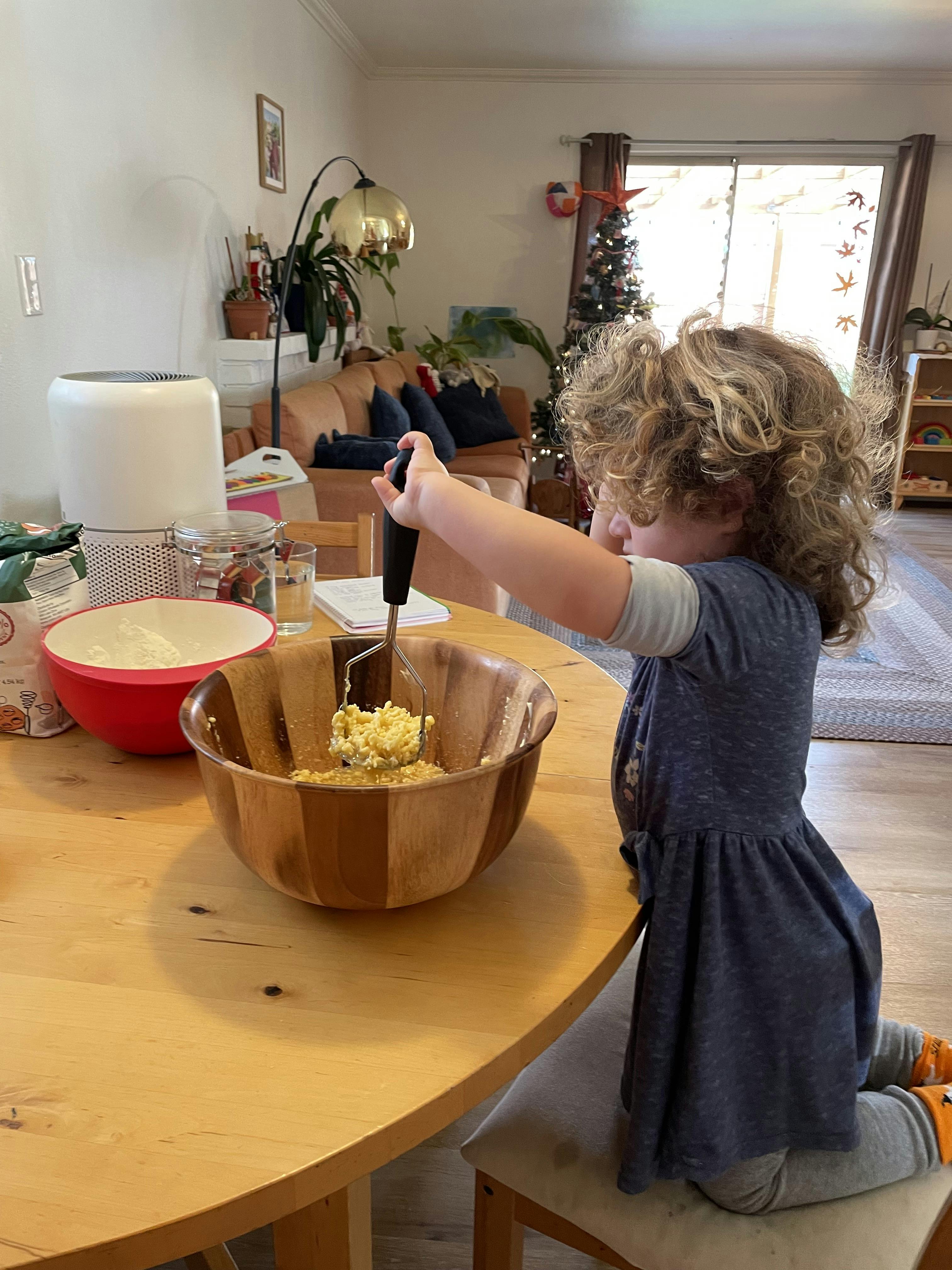 Child kneeling on a chair to mash food in a wooden bowl on a table