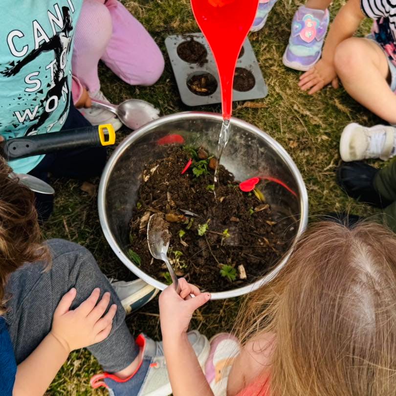 Children playing with dirt and water in a metal bowl outdoors