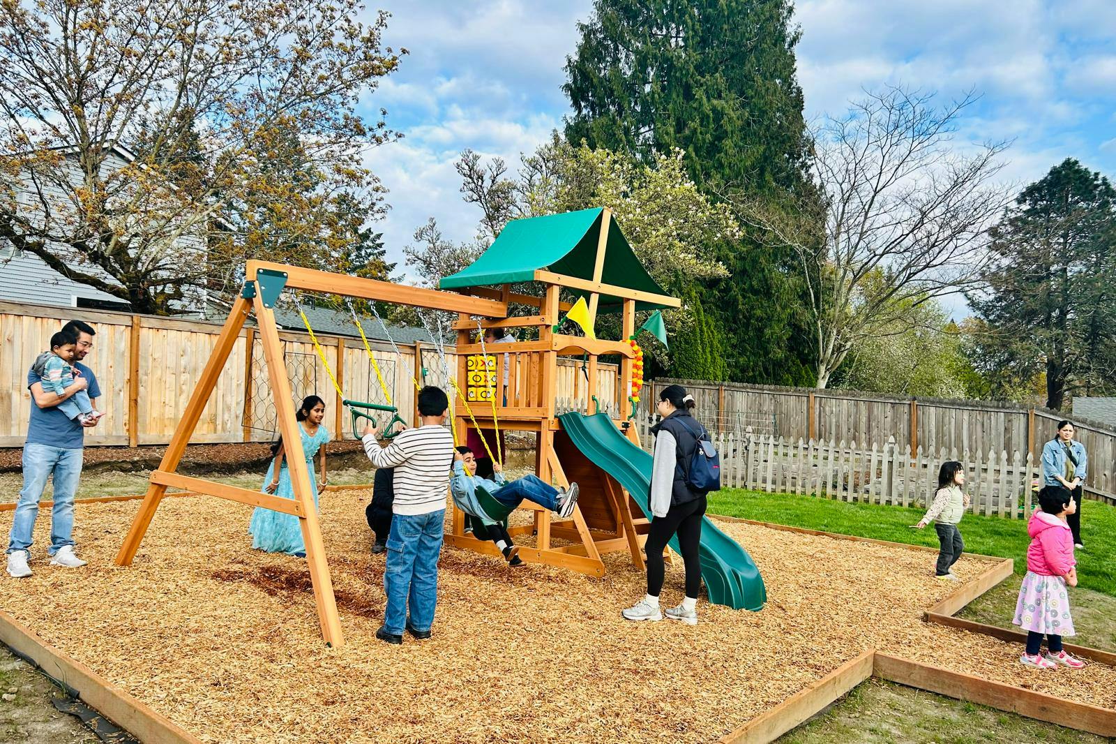 People and children playing on a wooden playground set with swings and slides