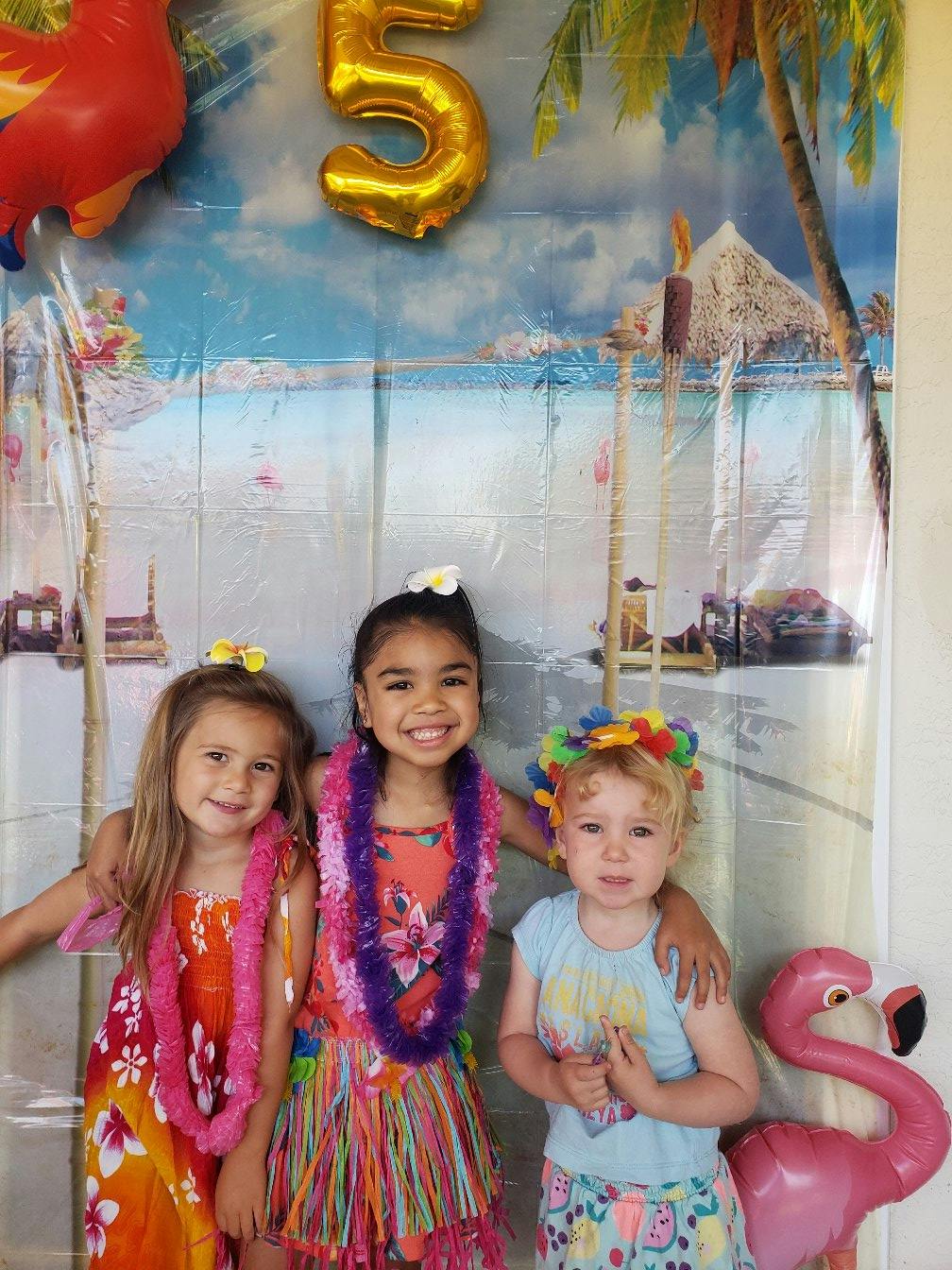 Three children in leis and tropical attire in front of a beach-themed backdrop