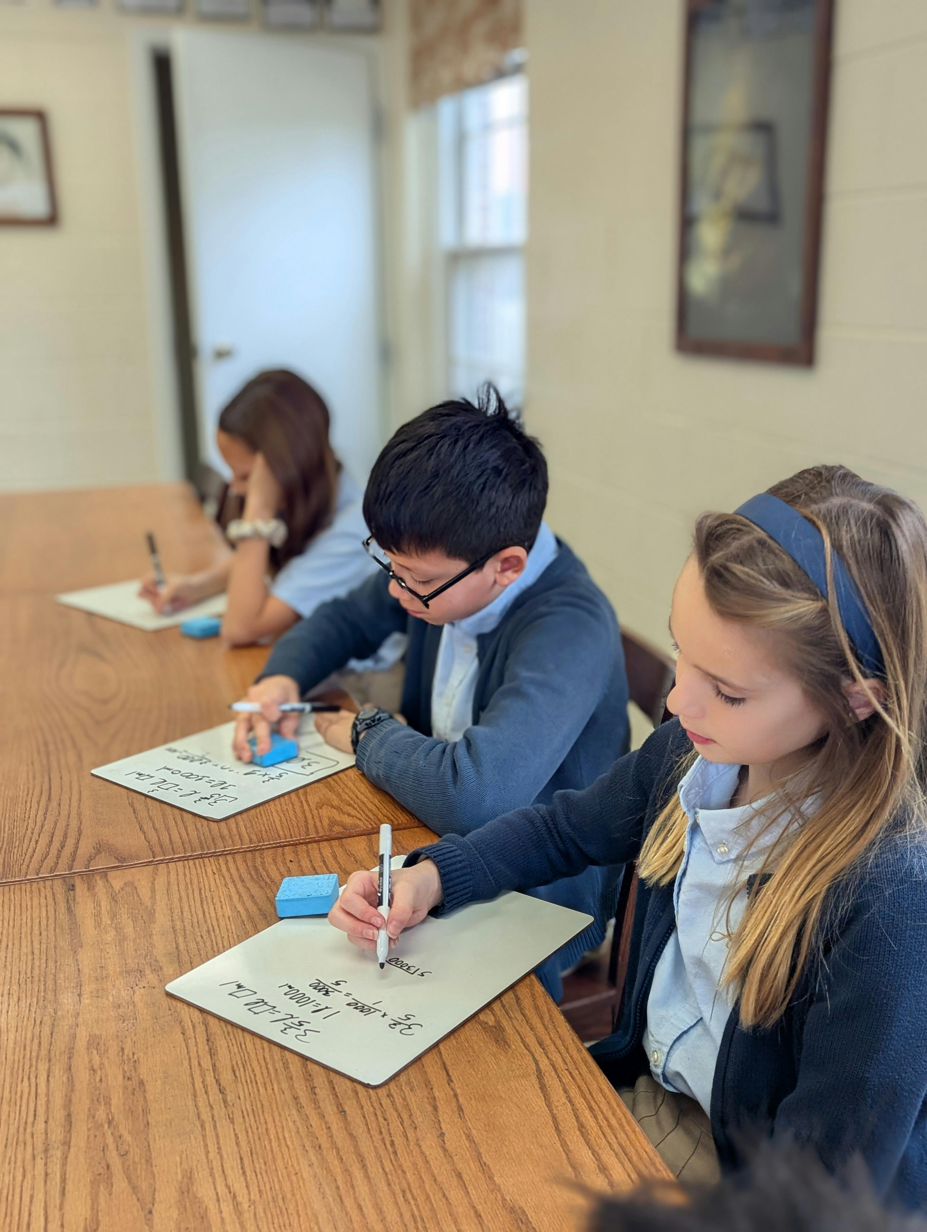 Children writing on whiteboards at a wooden table in a classroom