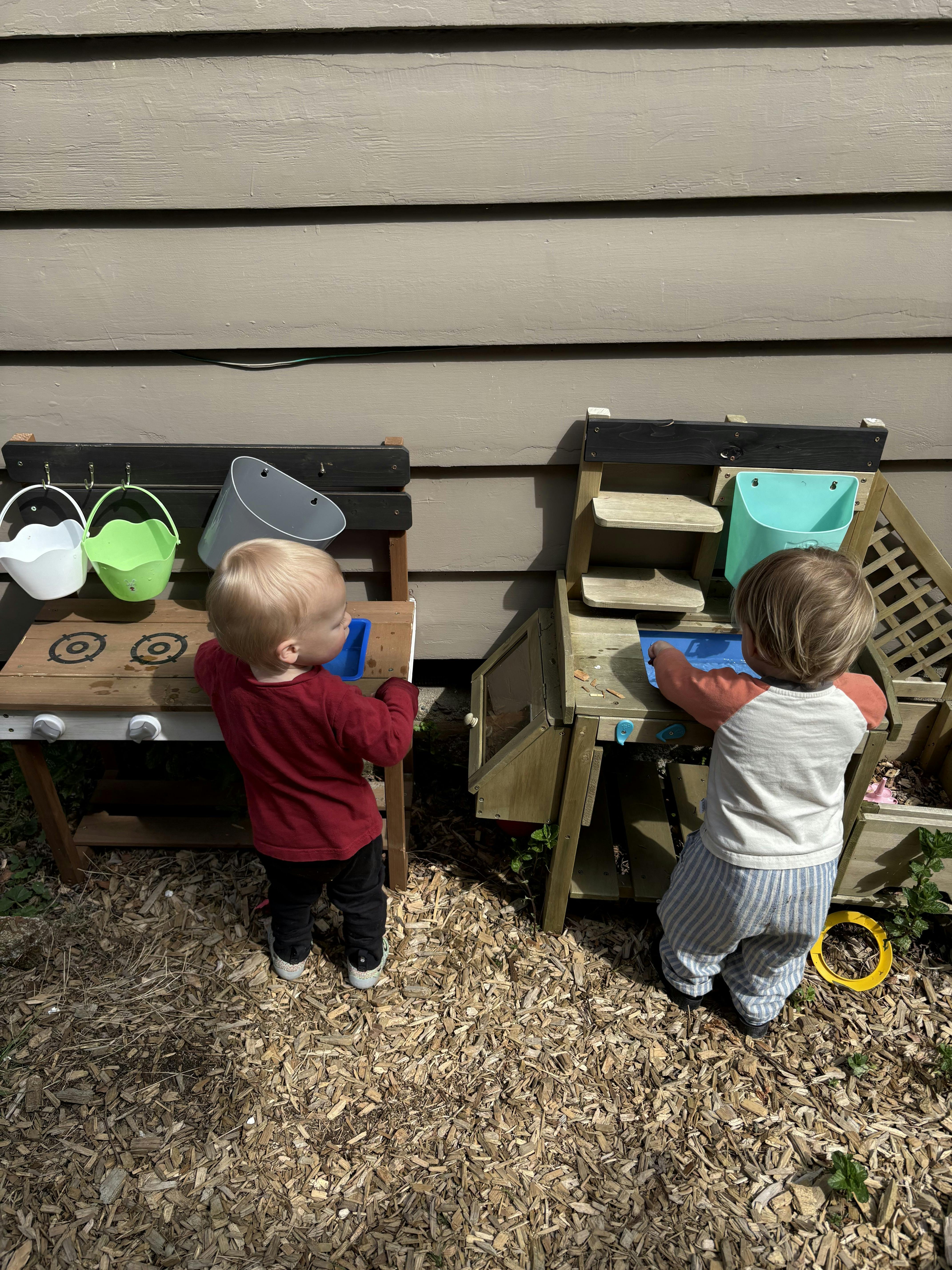 Two children playing at separate outdoor wooden play kitchens