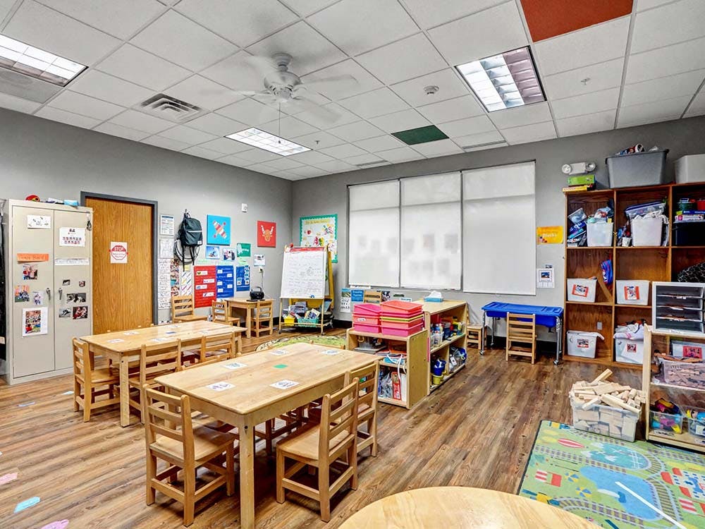 Classroom with small wooden tables and chairs, shelving units with supplies