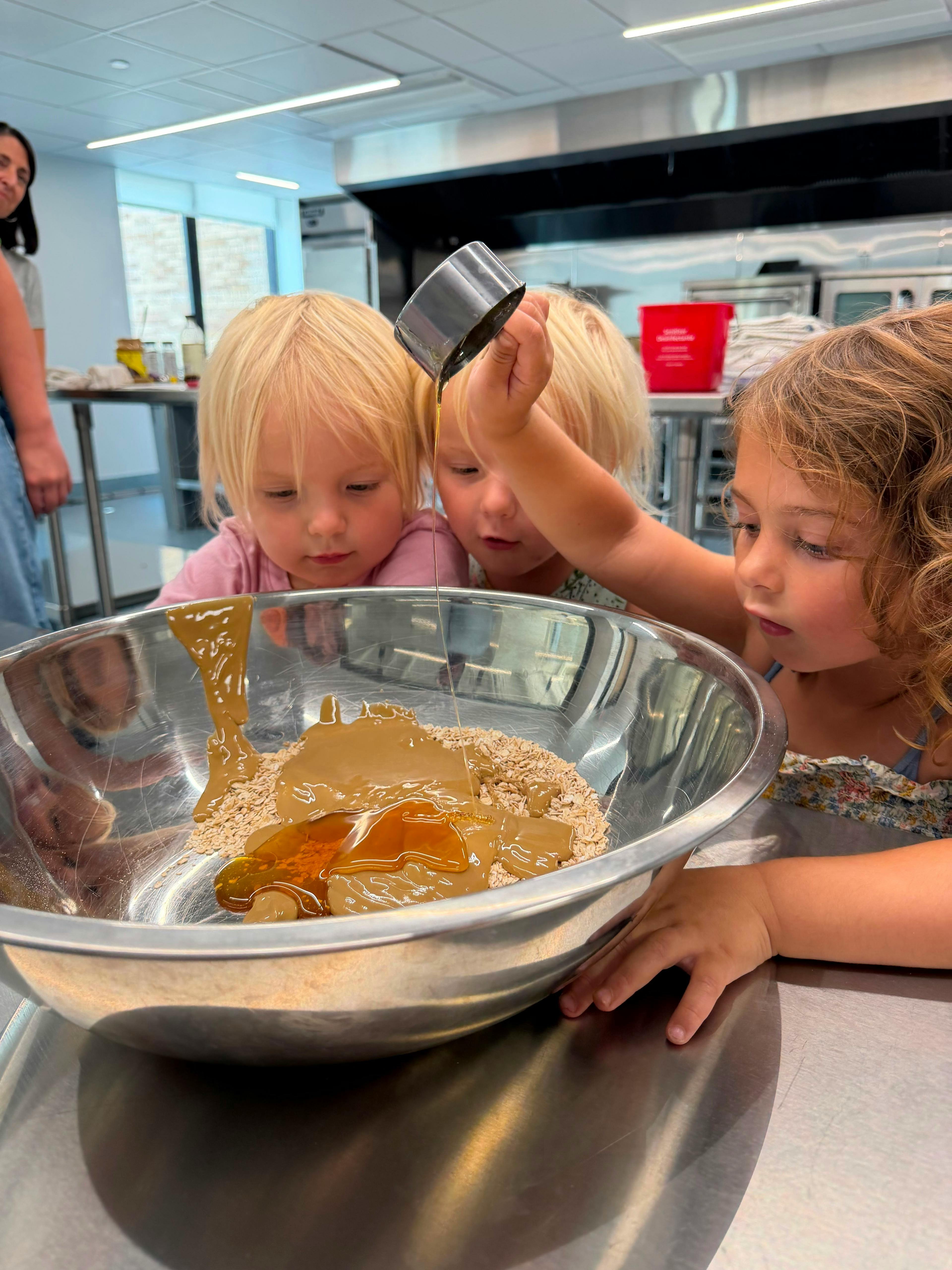 Three children pouring liquid into a bowl of oats in a kitchen