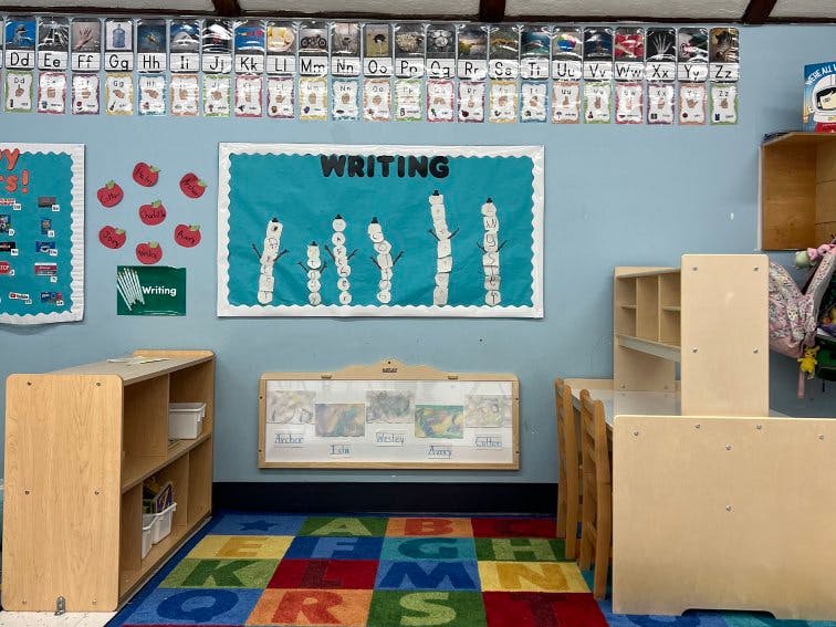 Classroom wall with alphabet charts, a writing bulletin board, and children's furniture