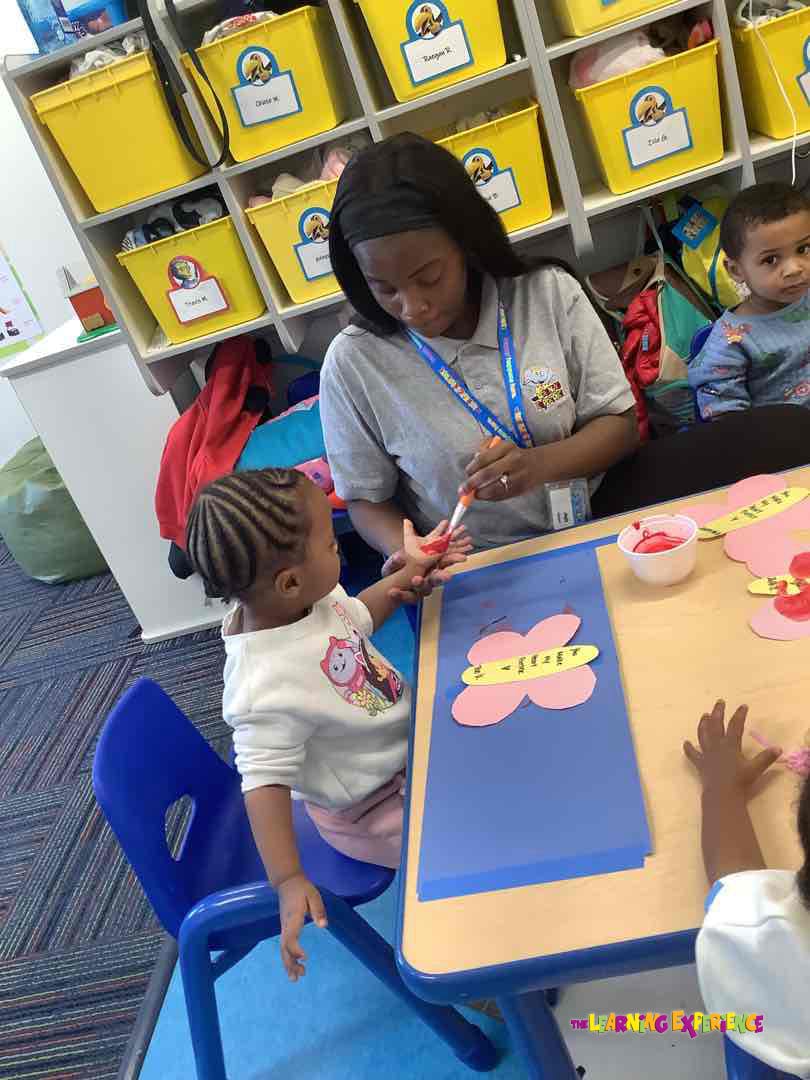 Adult helping a child with handprint painting at a table in a classroom