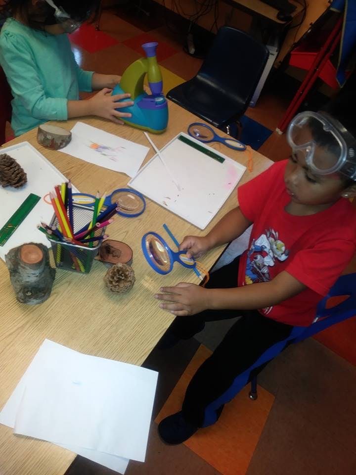 Child in safety goggles holding a magnifying glass at a table with another child