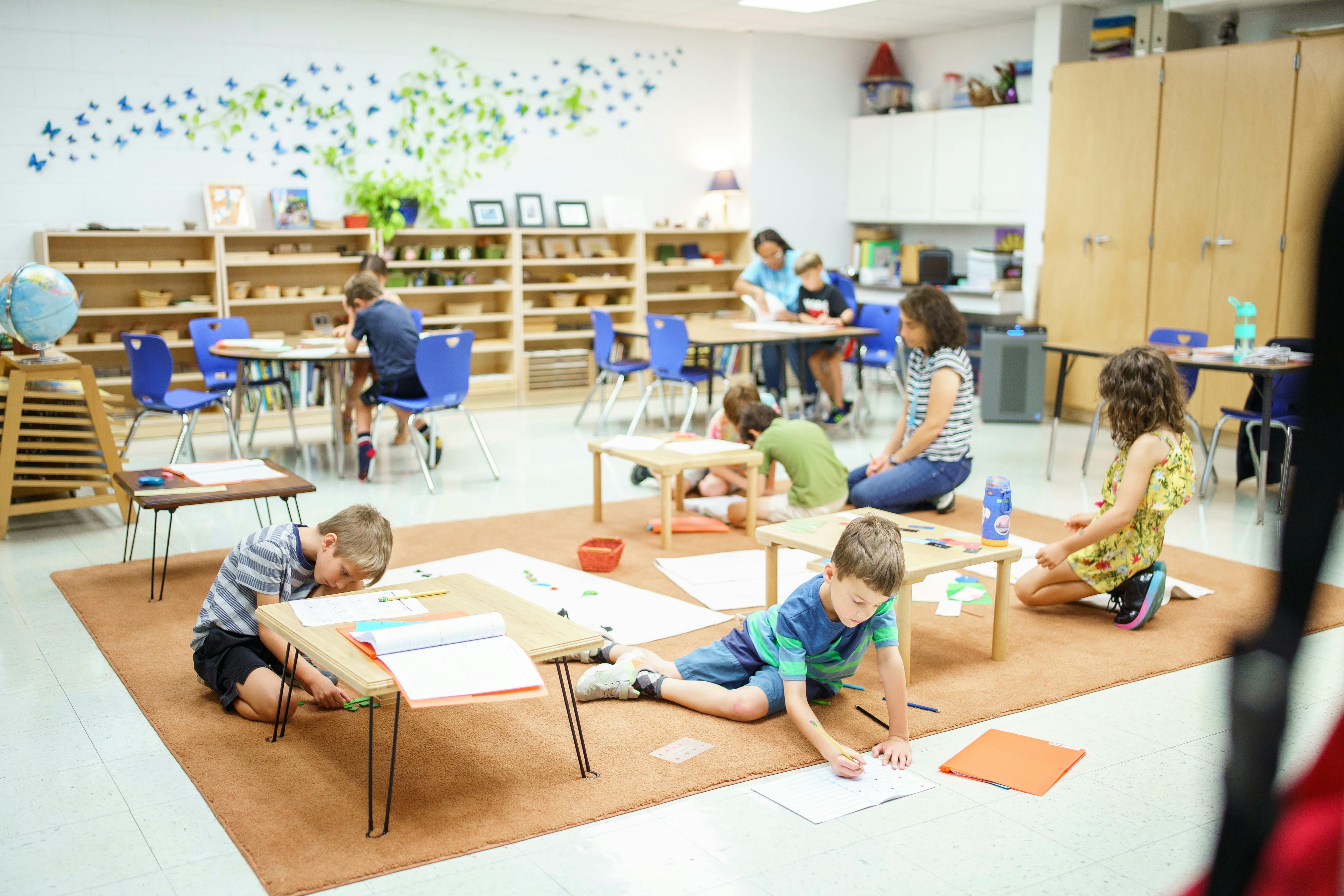 Children and adults in a classroom, some sitting at tables, others on the floor