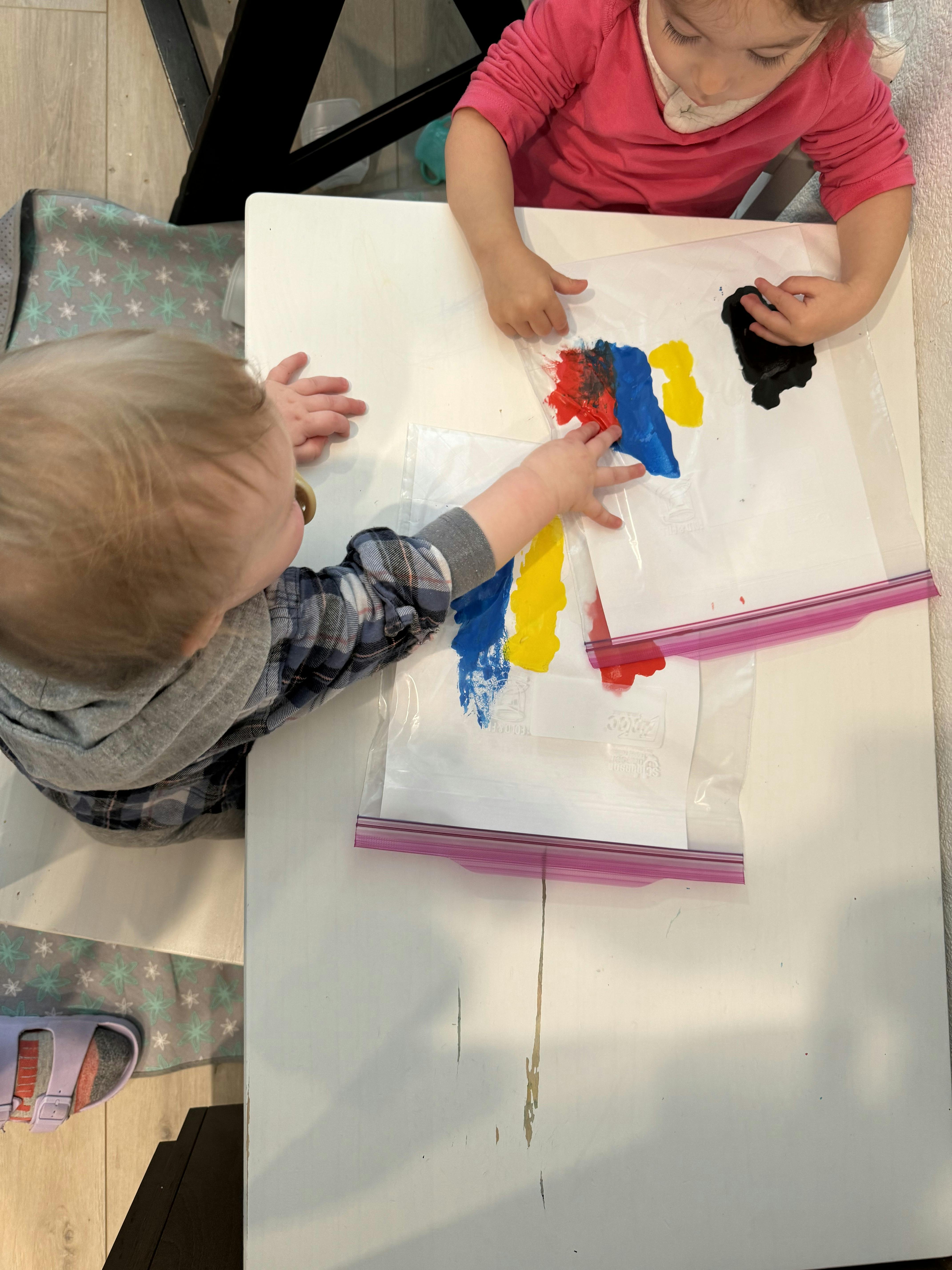Two children playing with paint inside plastic bags on a table