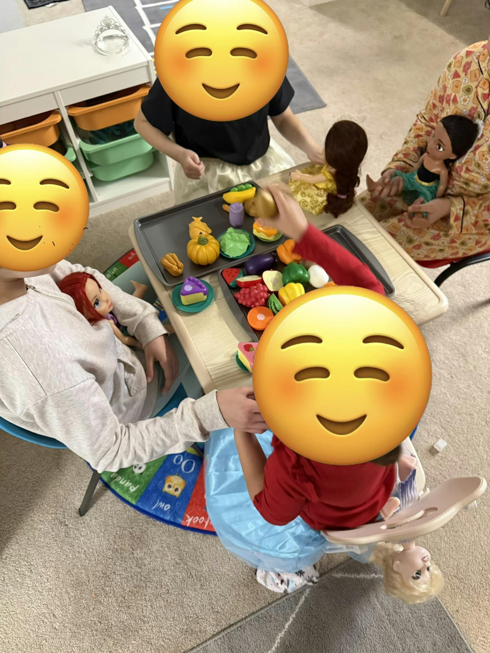 People playing with toy food at a small table, surrounded by dolls
