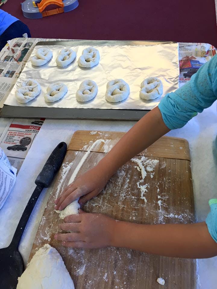 Child's hands rolling dough on a floured cutting board, pretzels on a baking sheet