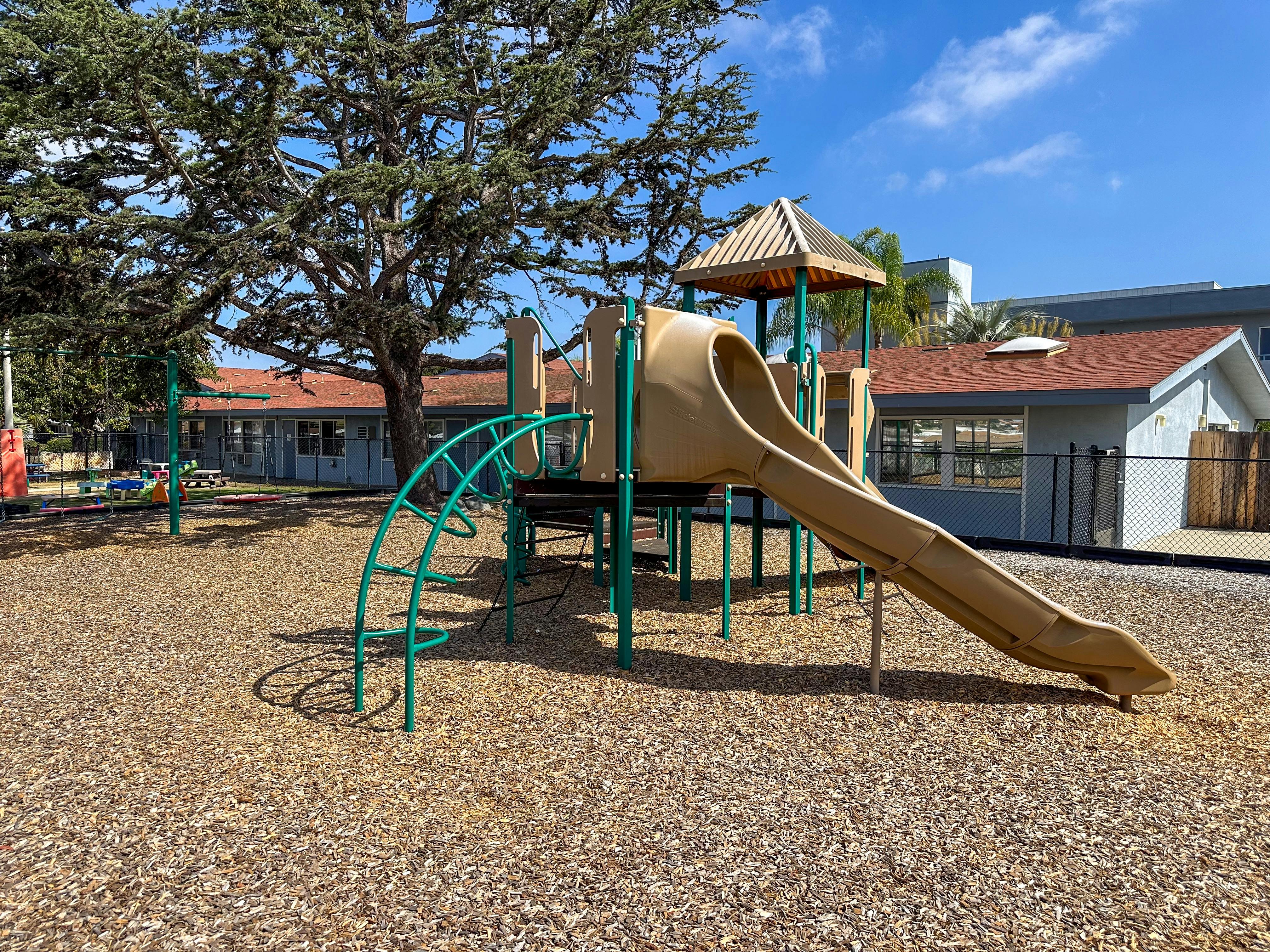 A tan and green playground slide and climber with wood chips on the ground