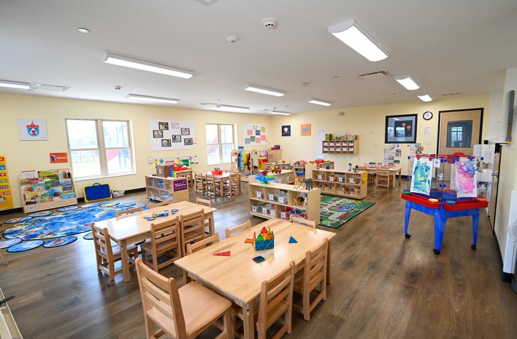 Classroom with multiple wooden tables and chairs, play mats, bookshelves, and art supplies