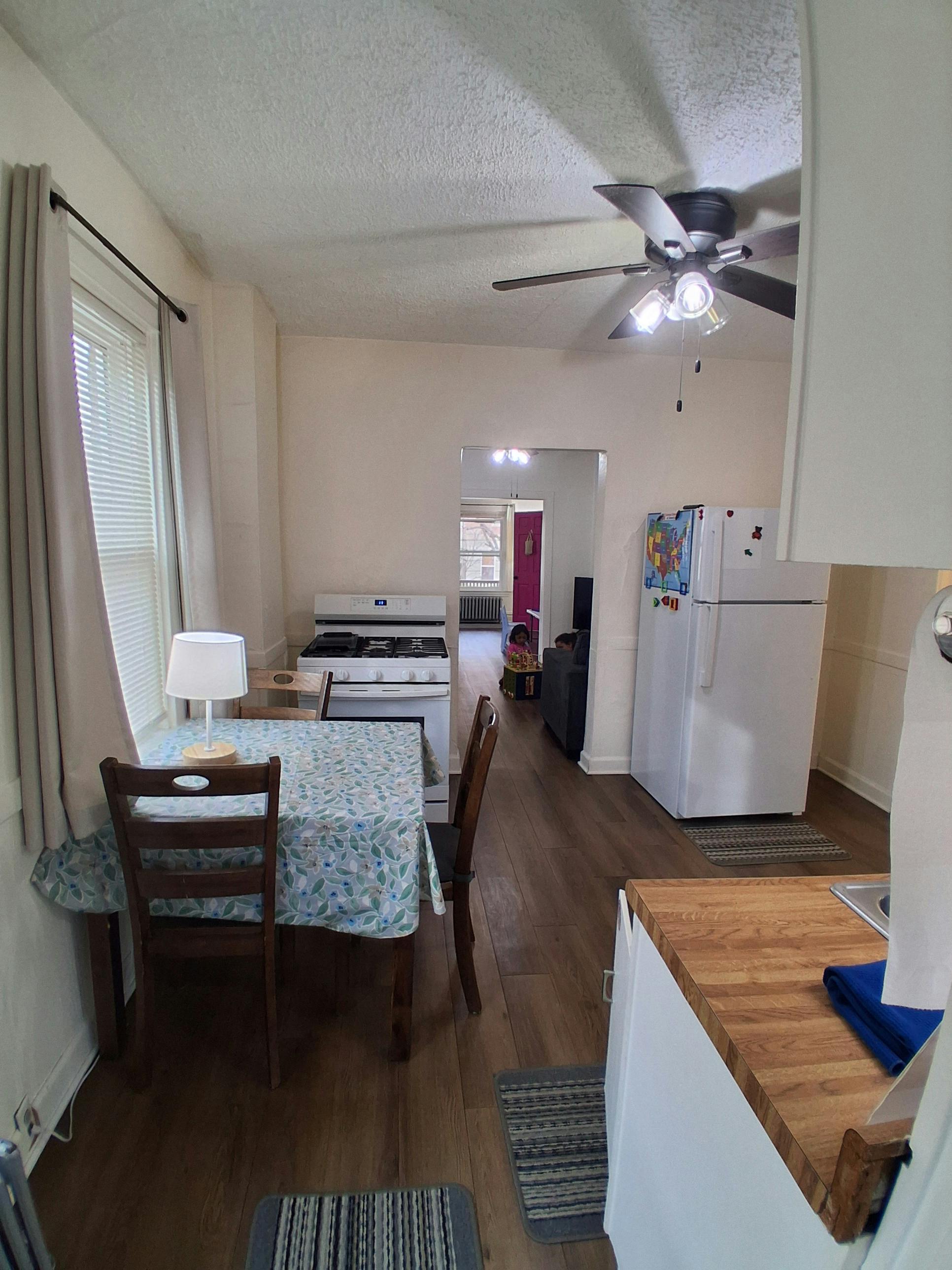 A small kitchen and dining area with a ceiling fan, table with patterned cloth, and white appliances