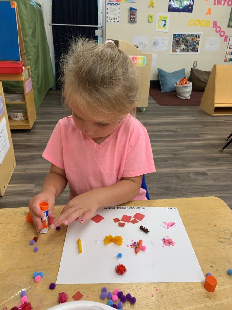 Child using a glue stick to attach craft items to paper in a classroom