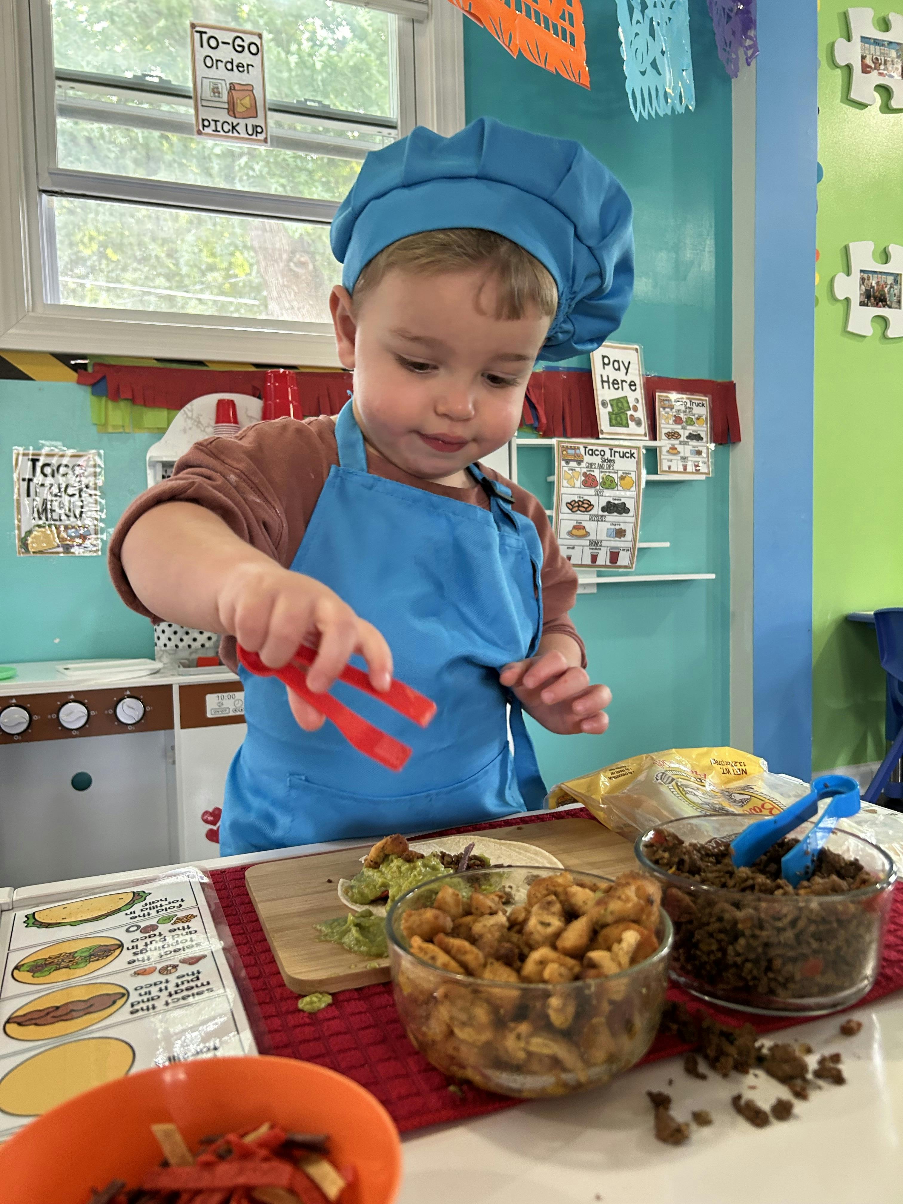 Child wearing a chef hat and apron using tongs to make a taco